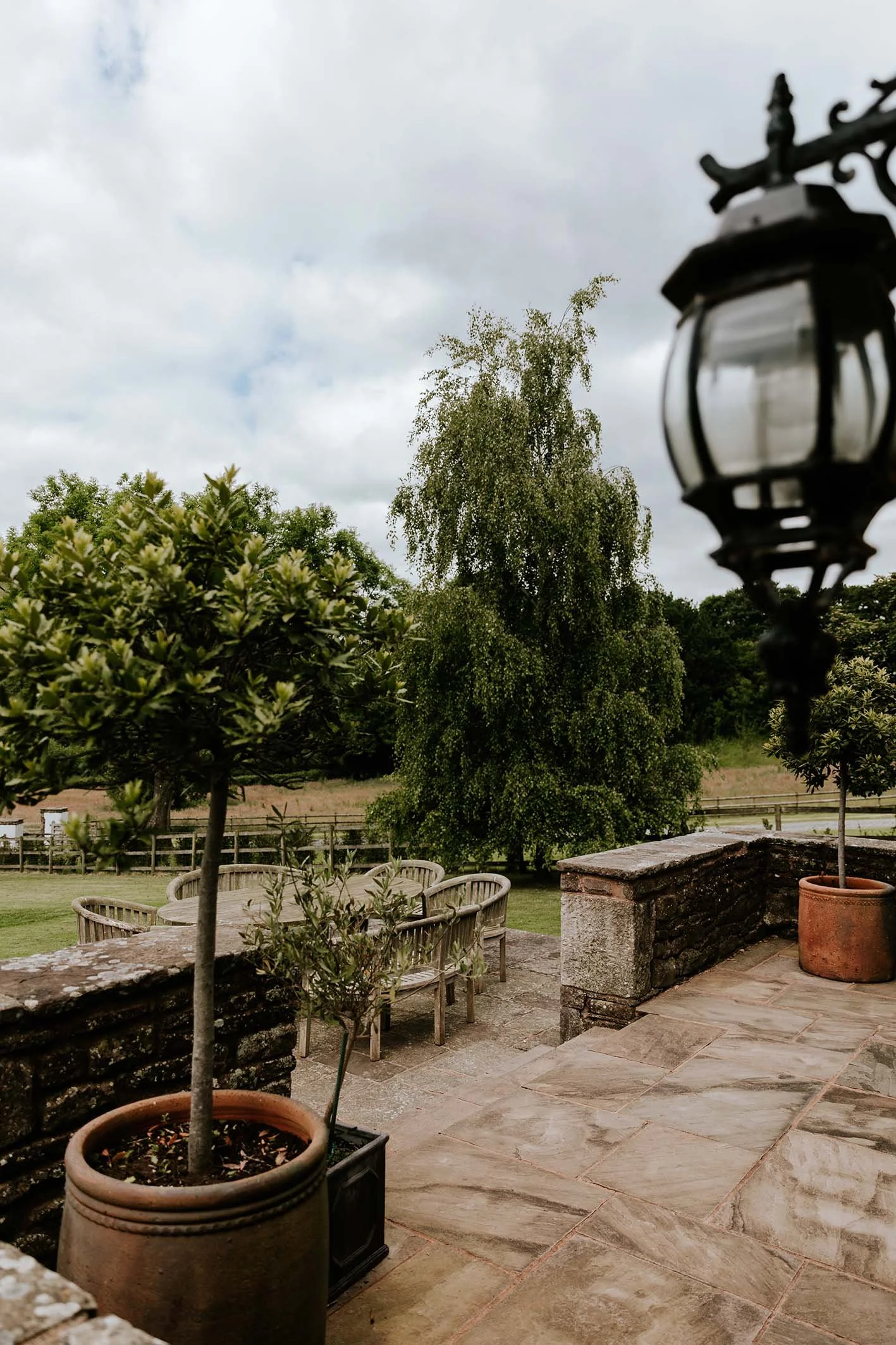 A stone patio with potted plants, wooden patio furniture, and large trees in a garden under cloudy sky.