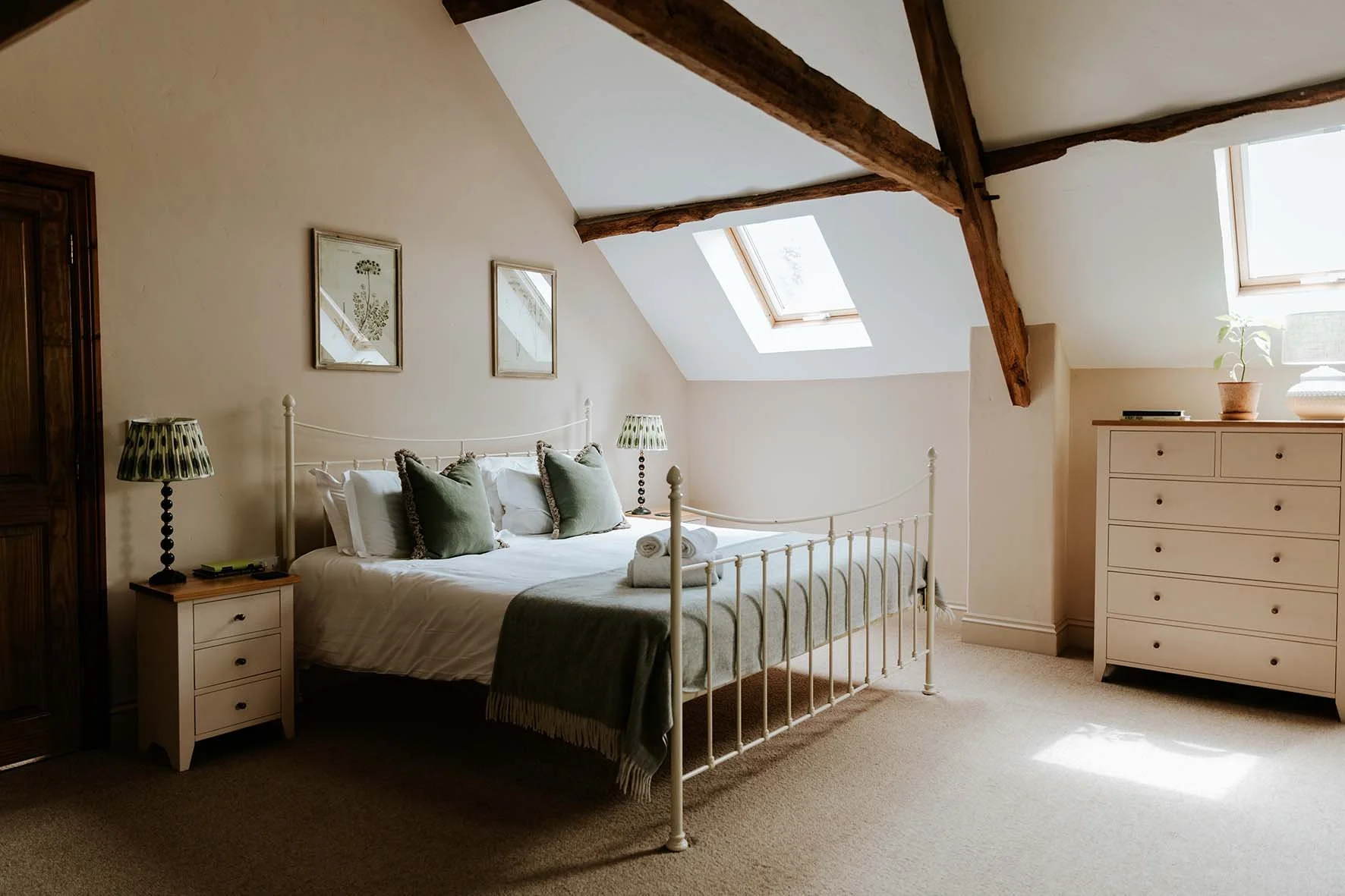A cozy bedroom with a white metal bed frame, topped with white and green pillows and a gray throw blanket, in an attic space with sloped ceiling and skylight windows.
