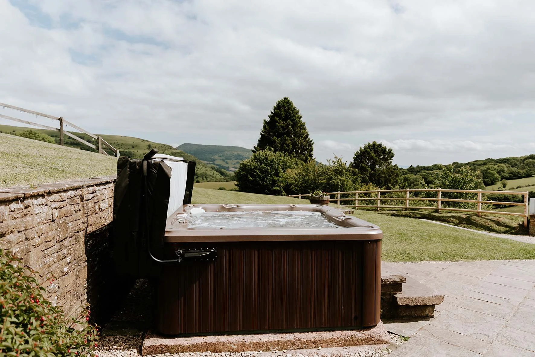 A hot tub with water on a patio outdoors, surrounded by a stone wall and a countryside landscape with rolling hills, trees, and a cloudy sky.