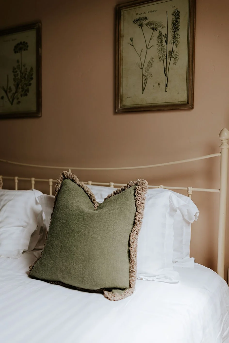A bedroom with a white bed, green pillow with a fuzzy border, and framed botanical prints on a pink wall.