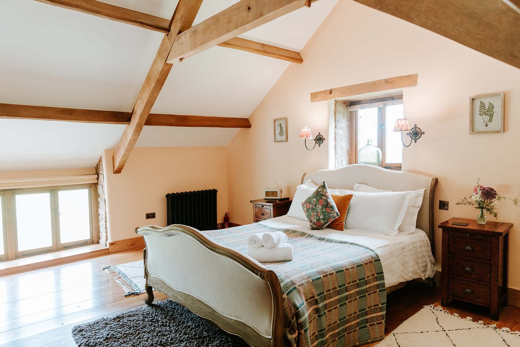 Cozy bedroom with a wooden ceiling, large bed with a white headboard, colorful pillows, and rolled towels. Natural light from a window, with wooden nightstands, a small vase with flowers, and wall art.