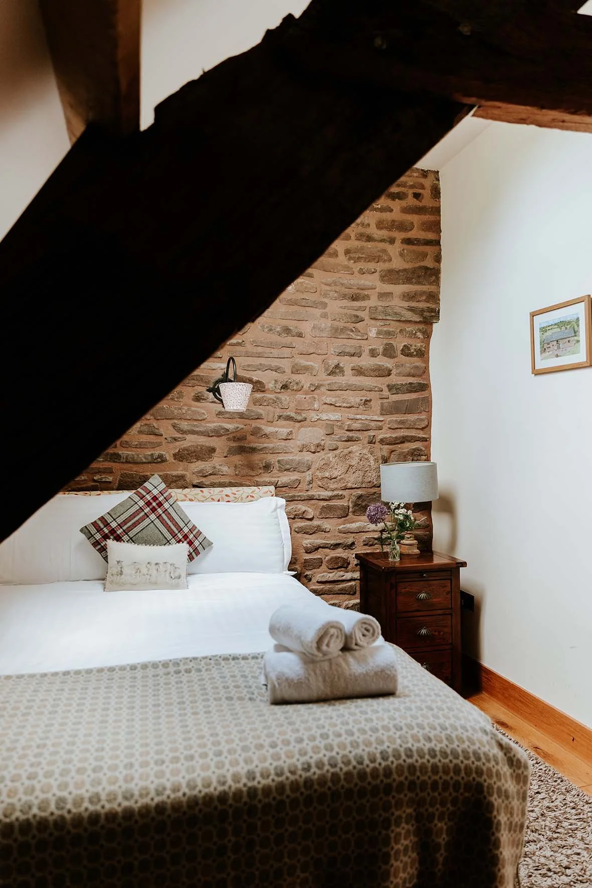 A cozy bedroom with a white bed, decorative pillows, and neatly rolled towels on the bed. The room has a rustic brick wall, a wooden nightstand with a lamp and a vase of flowers, and a framed picture on the wall. A dark wooden beam partially obscures