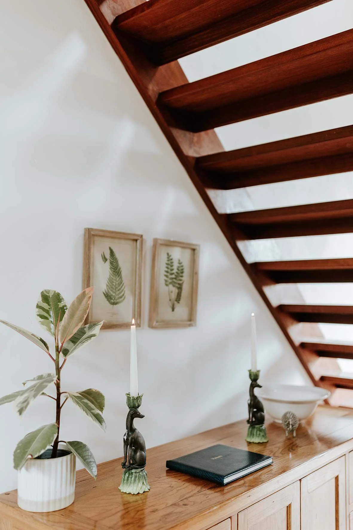 Wooden sideboard under a staircase with plants, candles, a black book, and decorative items, with framed botanical prints on the wall.