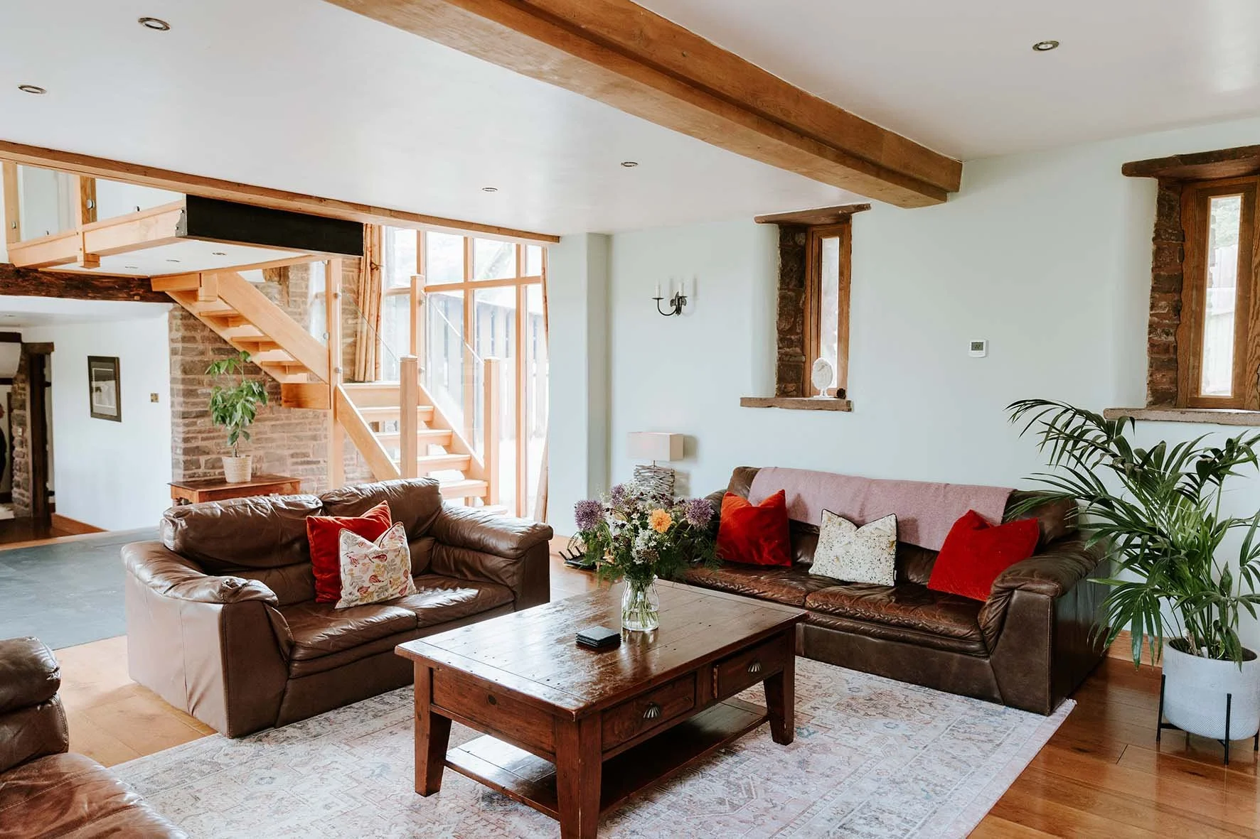 Living room with two brown leather sofas, a wooden coffee table with a vase of flowers, and large windows with wooden framing, exposing a staircase.