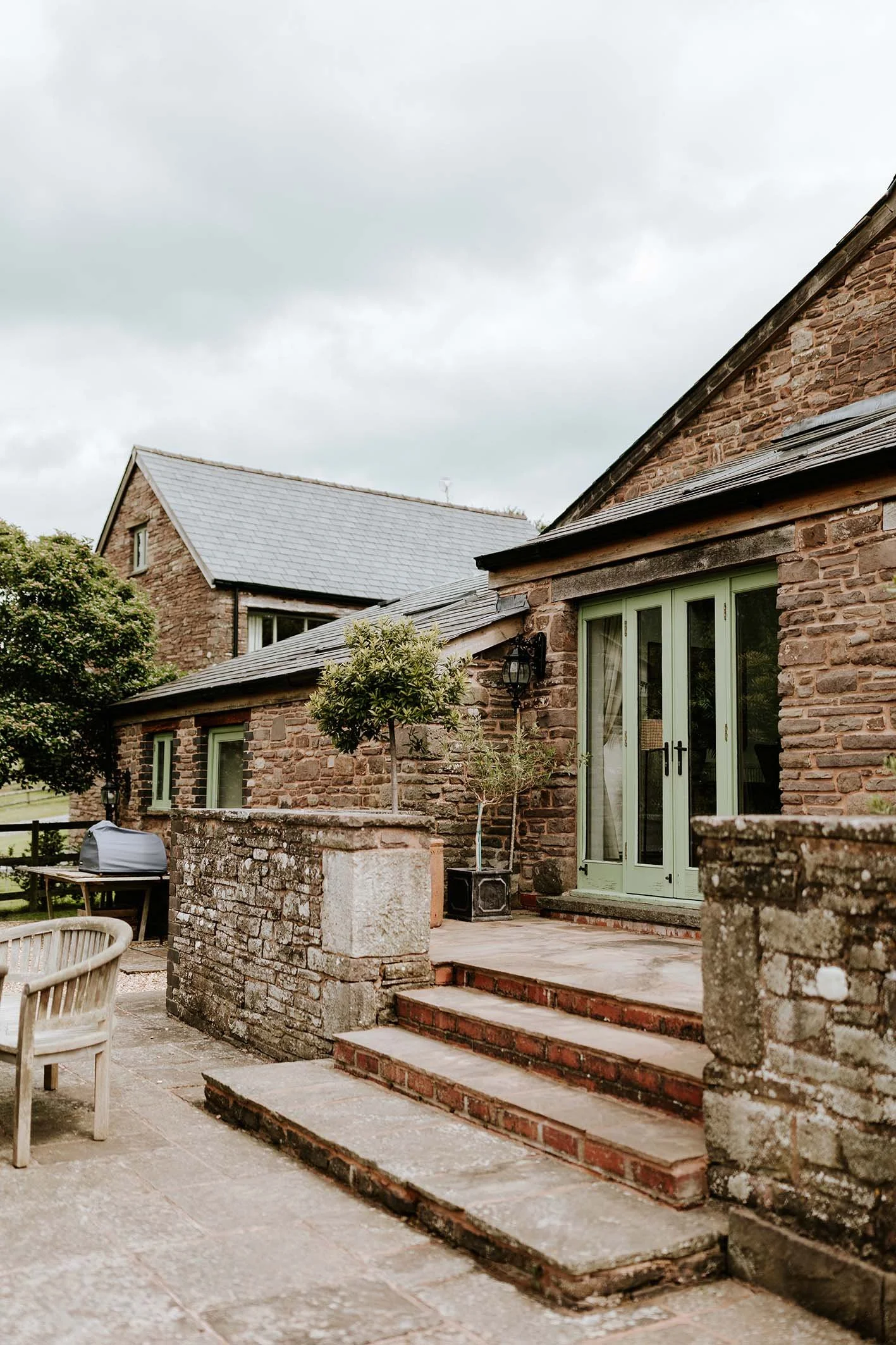A backyard patio area with stone steps leading to brick and stone house with green double doors, potted plants, and outdoor furniture against a cloudy sky.
