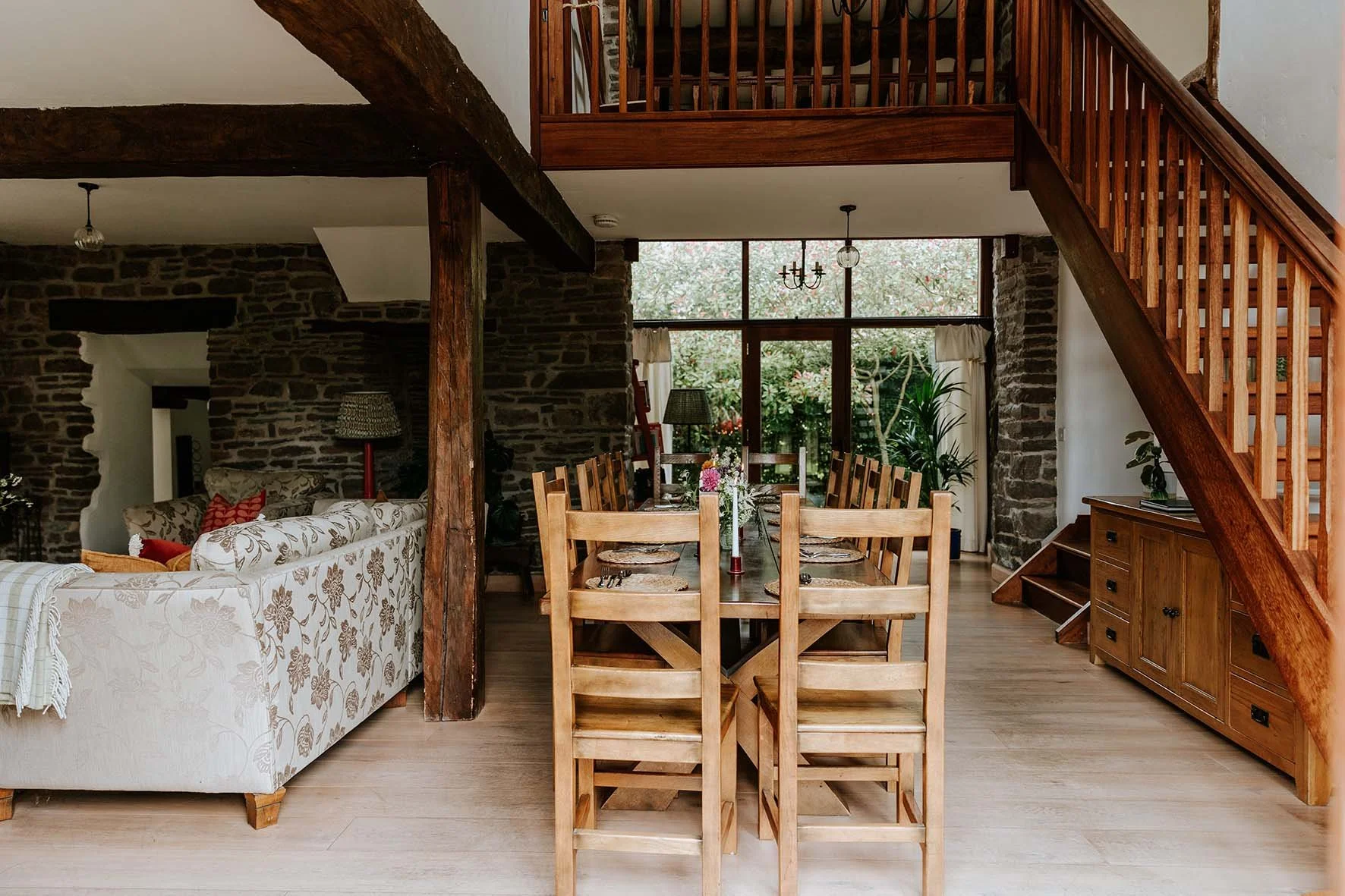 A rustic living and dining room with wooden furniture, exposed dark wood beams, and stone walls. There is a long wooden dining table with eight chairs, and an adjacent living space with a floral-patterned sofa. Large windows with curtains reveal gree