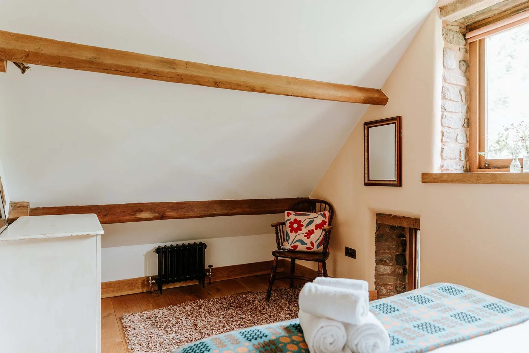 A cozy bedroom with a sloped ceiling, wooden beams, a window with a wooden frame, a small mirror, a chair with a floral cushion, a stack of rolled towels, a small white cabinet, a plush rug, and a bed with a colorful patterned blanket.