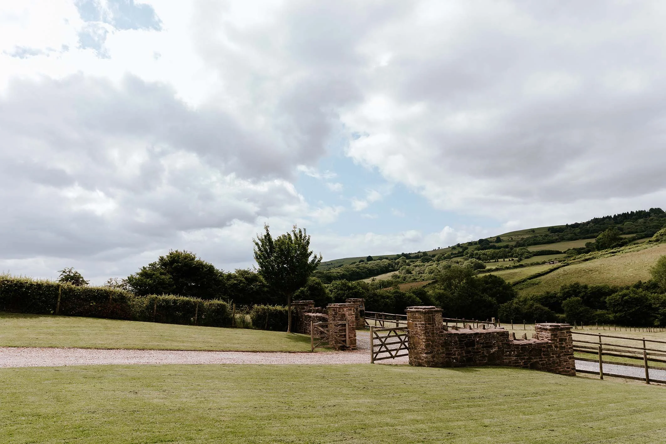 A rural landscape with a grassy lawn, a stone wall with wooden gates, hedges, trees, rolling green hills, and a partly cloudy sky.