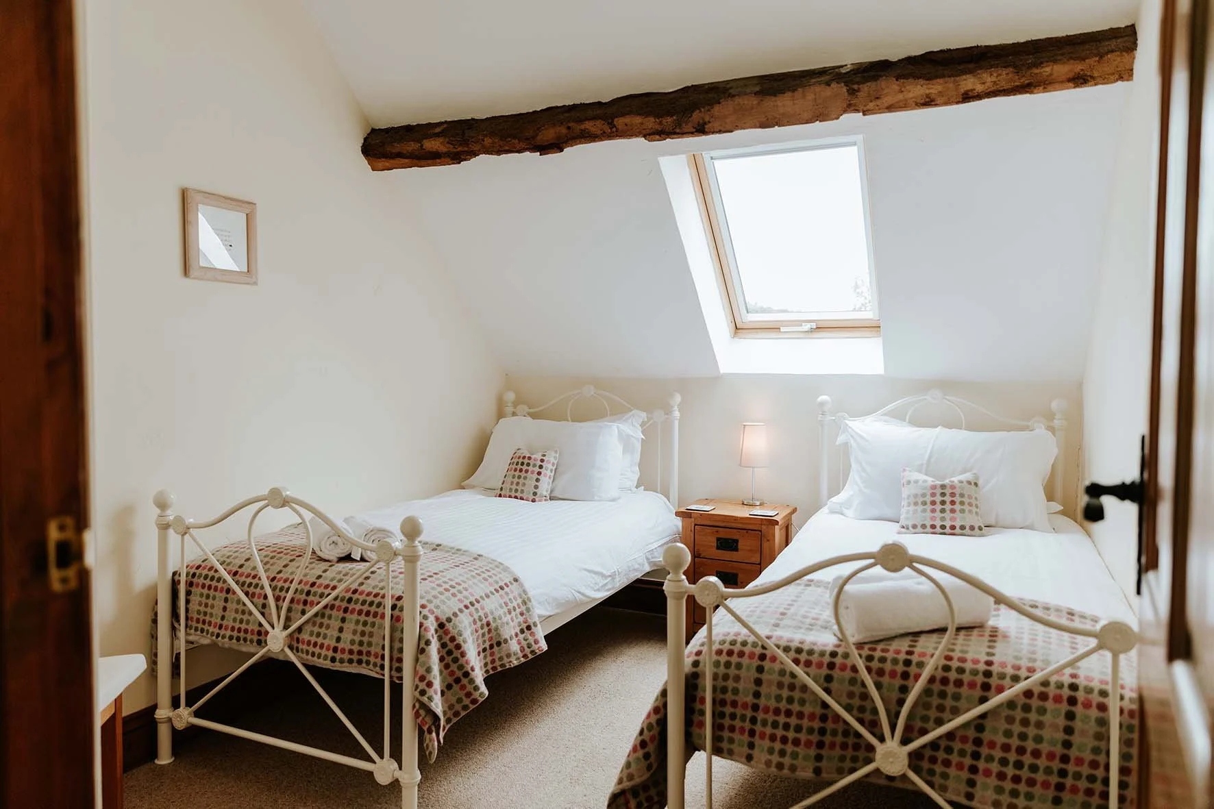 A cozy bedroom with two white metal beds with dotted bedspreads, a wooden nightstand with a lamp between the beds, and a dormer window on the slanted ceiling allowing natural light.