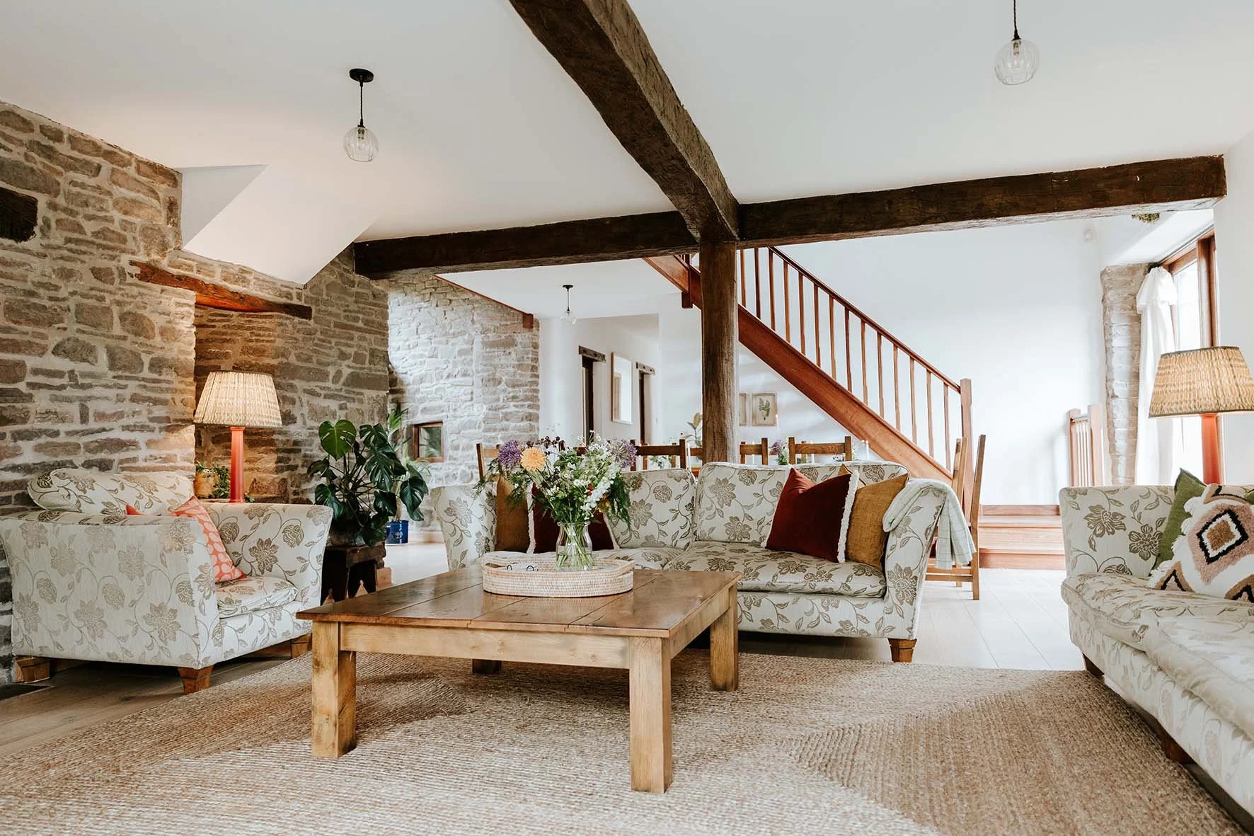 Living room with rustic stone walls, wooden beams, floral patterned sofas, a wooden coffee table with a flower vase, and large windows with curtains.