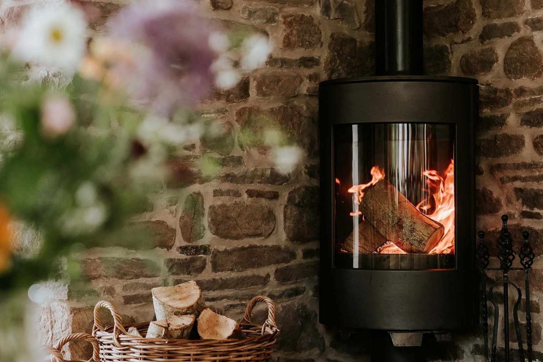 A wood-burning stove with burning logs against a brick wall, with a basket of chopped wood in front.