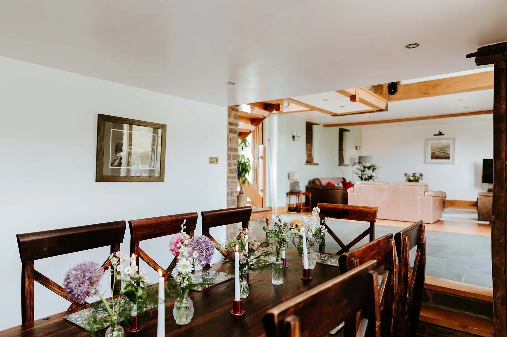 Dining room with a wooden table decorated with flowers in glass vases and white candles, set in a home with wooden accents, a staircase, and a living area with sofas and plants in the background.