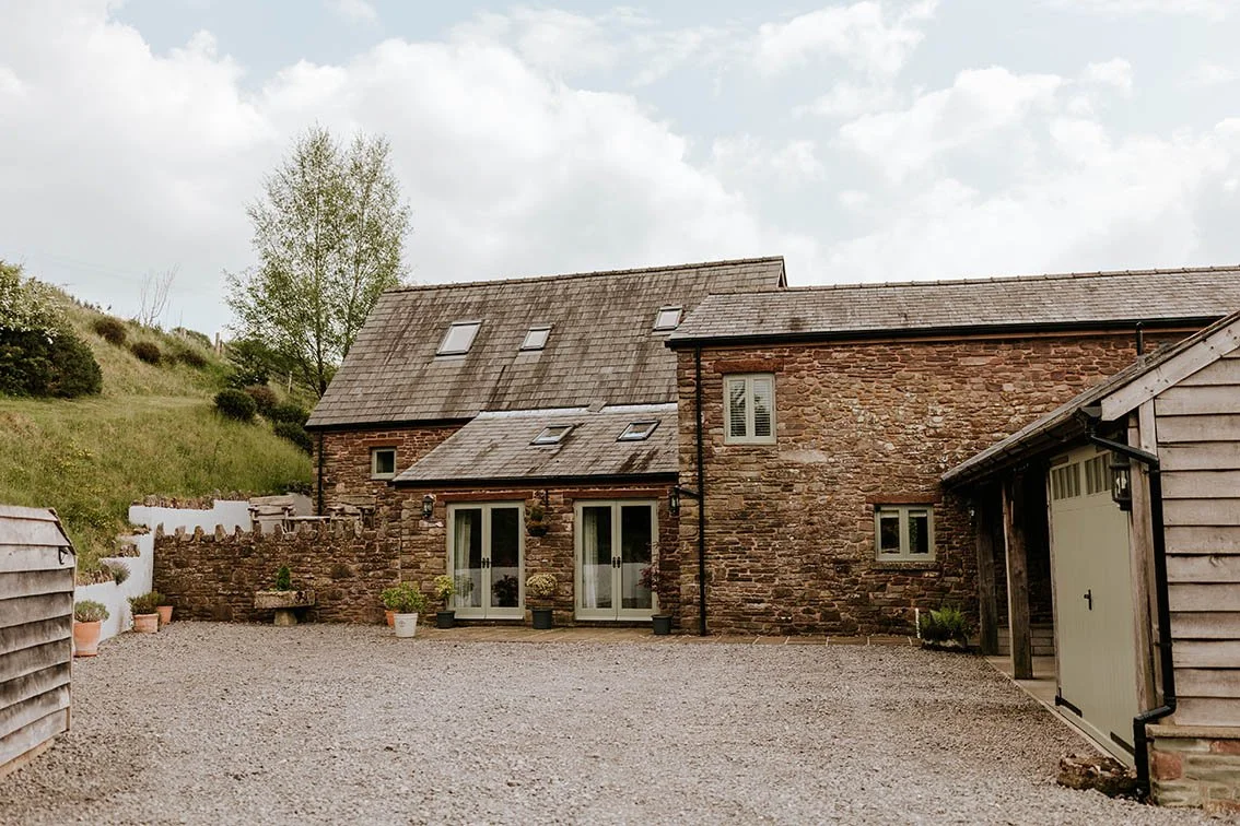 Stone house with multiple small windows and glass doors, gravel driveway, wooden shed, potted plants, and a hillside with grass and trees in the background.