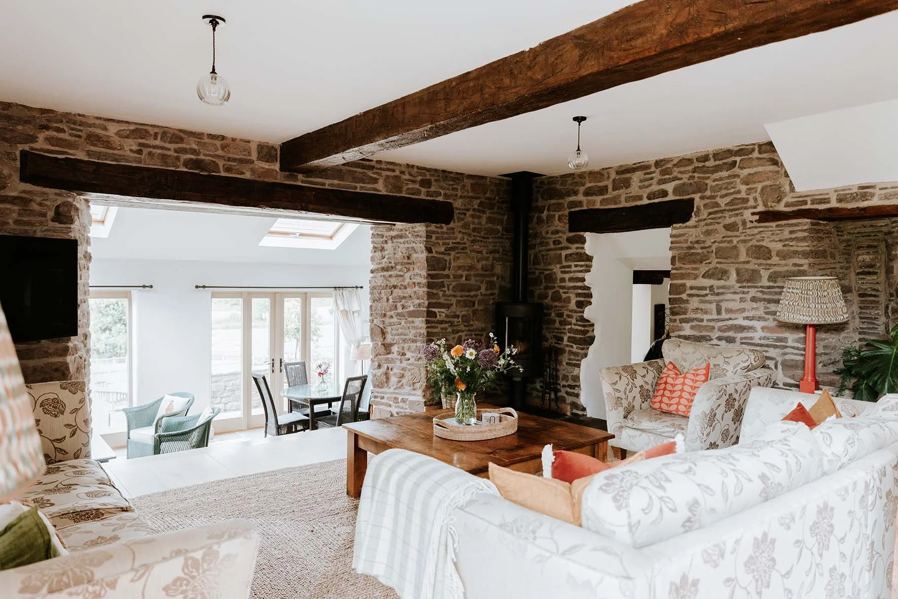 Living room with exposed brick walls, wooden beams, a fireplace, floral-patterned sofas, colorful pillows, a wooden coffee table with a bouquet of flowers, a standing lamp, and large windows with curtains leading to a dining area with a table and cha