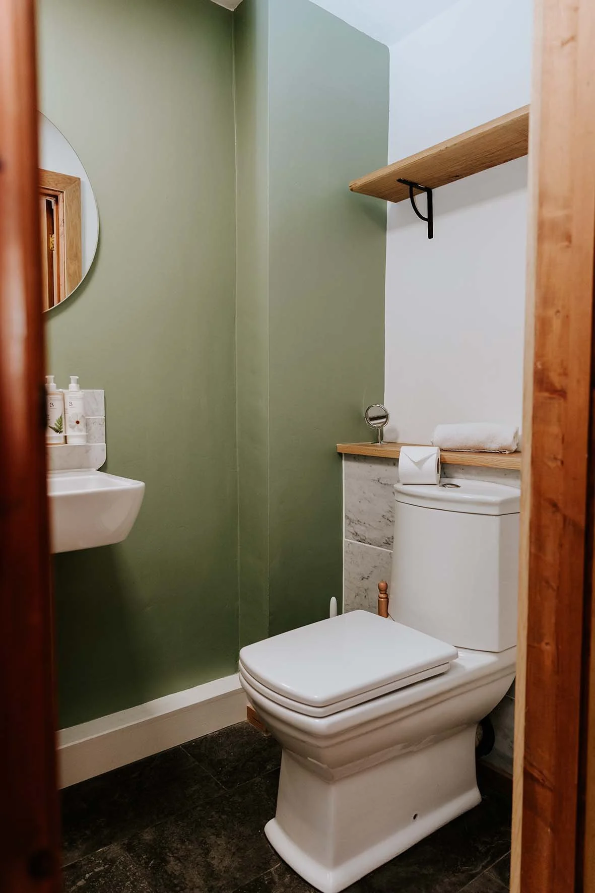 Small bathroom with green and white walls, a white toilet with a wooden pipe, a small white sink, a mirror, a wooden shelf with a rolled towel and toiletries, and a mirror reflecting wood paneling.