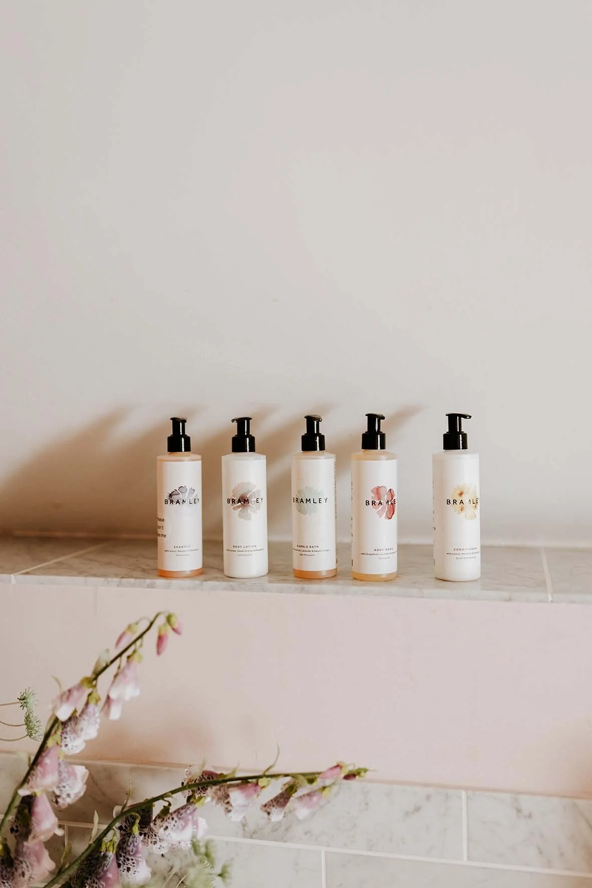 Five white bottles with black pumps labeled 'Bramley' on a marble shelf, with a blurred pink flowering plant in the foreground.