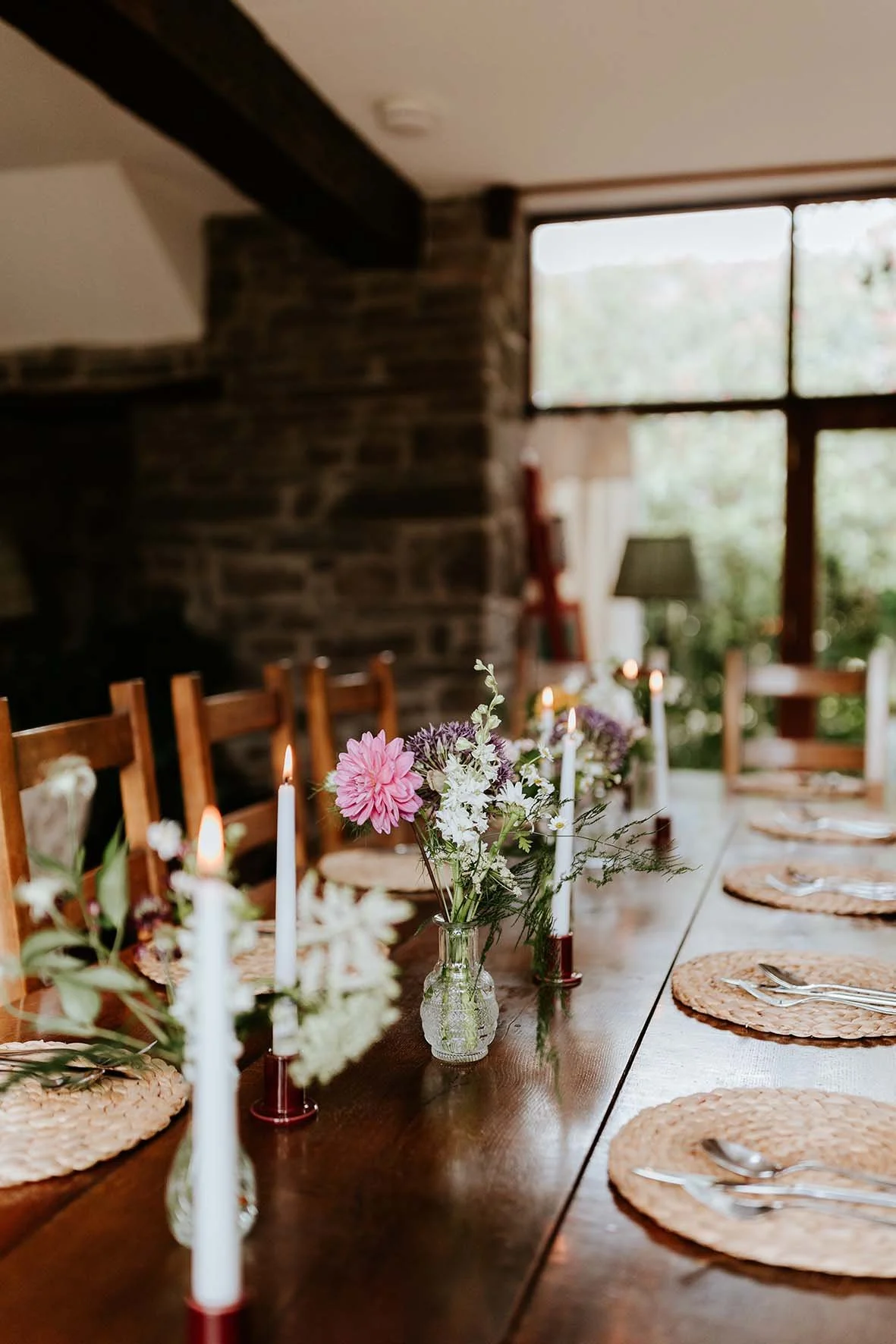 A long wooden dining table set for a meal, with woven placemats, silverware, white candles in holders, and small glass vases with pink and white flowers.