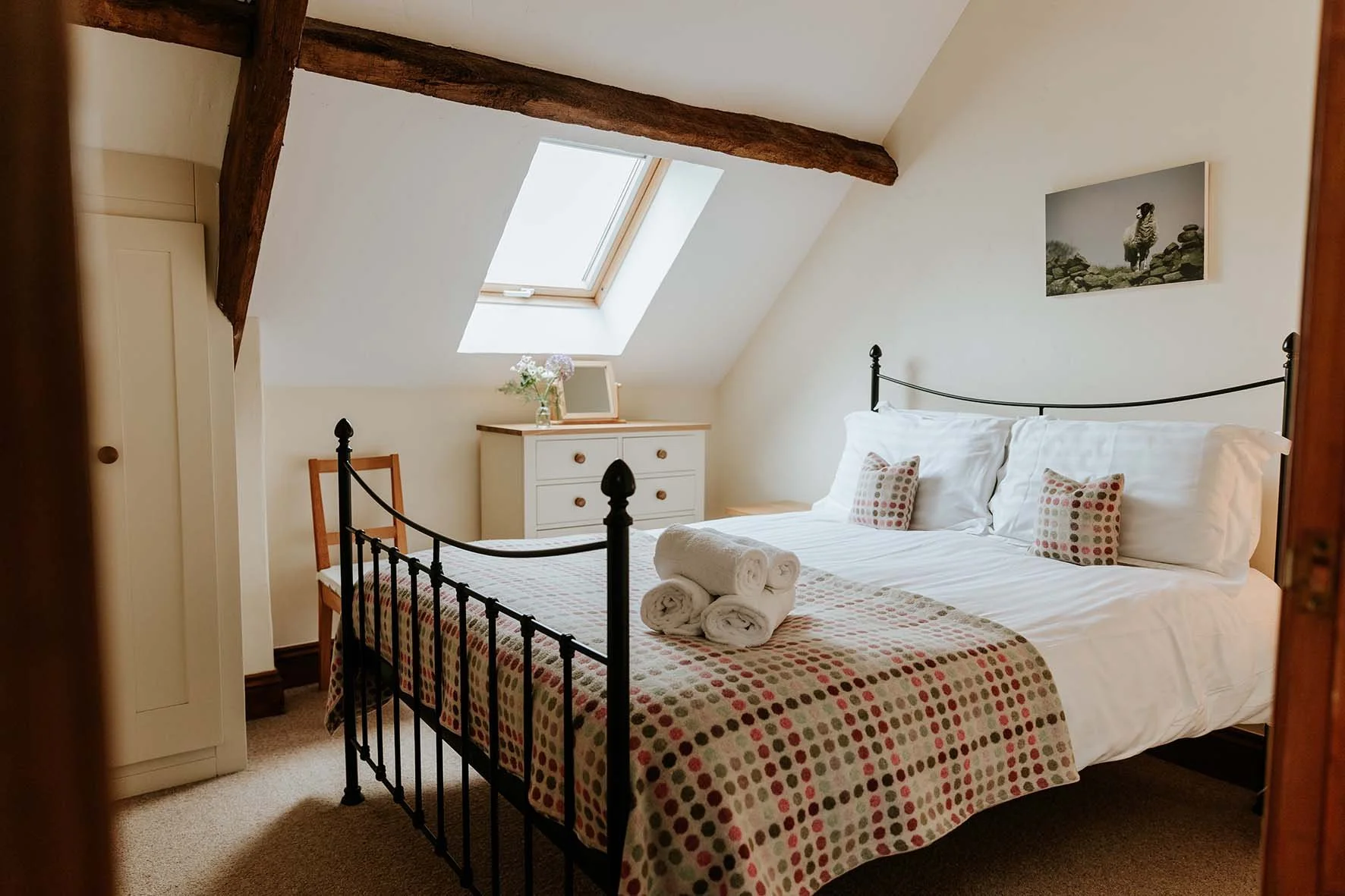 A cozy bedroom with a sloped ceiling, skylight window, white bedding, patterned bed runner, and rolled towels on the bed. There is a white dresser with a vase of flowers and a picture frame, a wooden chair, a wardrobe, and a framed photo of a dog on 