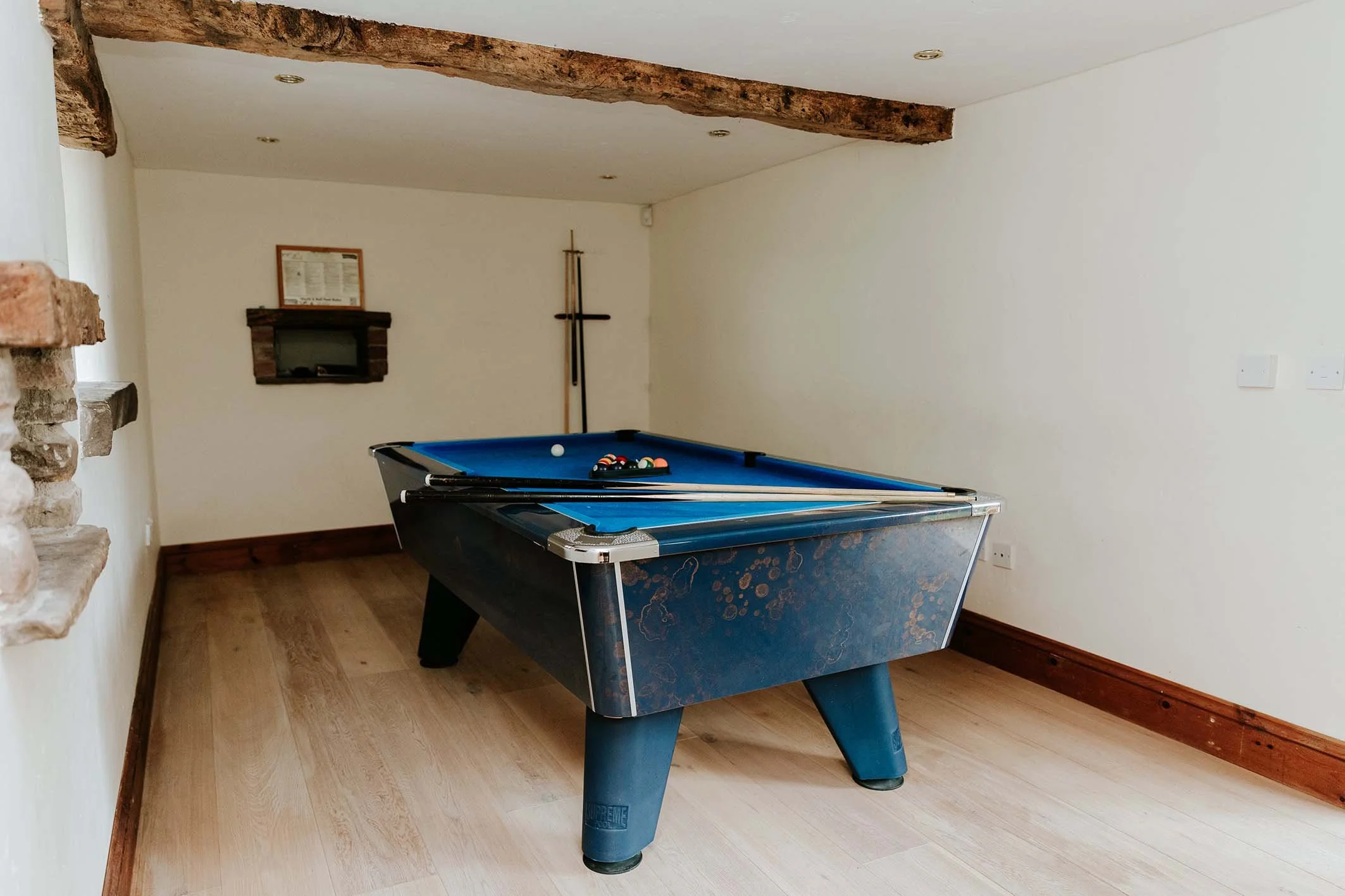 A blue pool table with pool balls and cues inside a room with white walls, wooden floor, exposed wooden beams, and a small fire place on the wall.