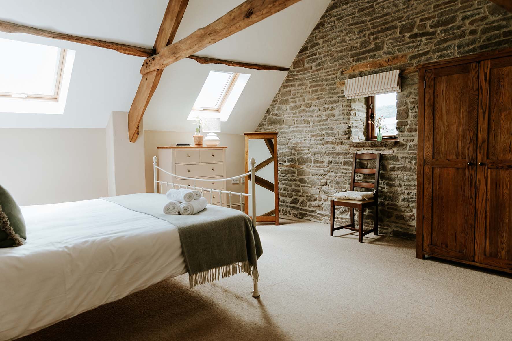 A cozy bedroom with a sloped ceiling and exposed wooden beams, featuring a bed with white linens and rolled towels, a beige dresser, a wooden chair, a large wooden wardrobe, and a small window on a stone wall, illuminated by two skylights.