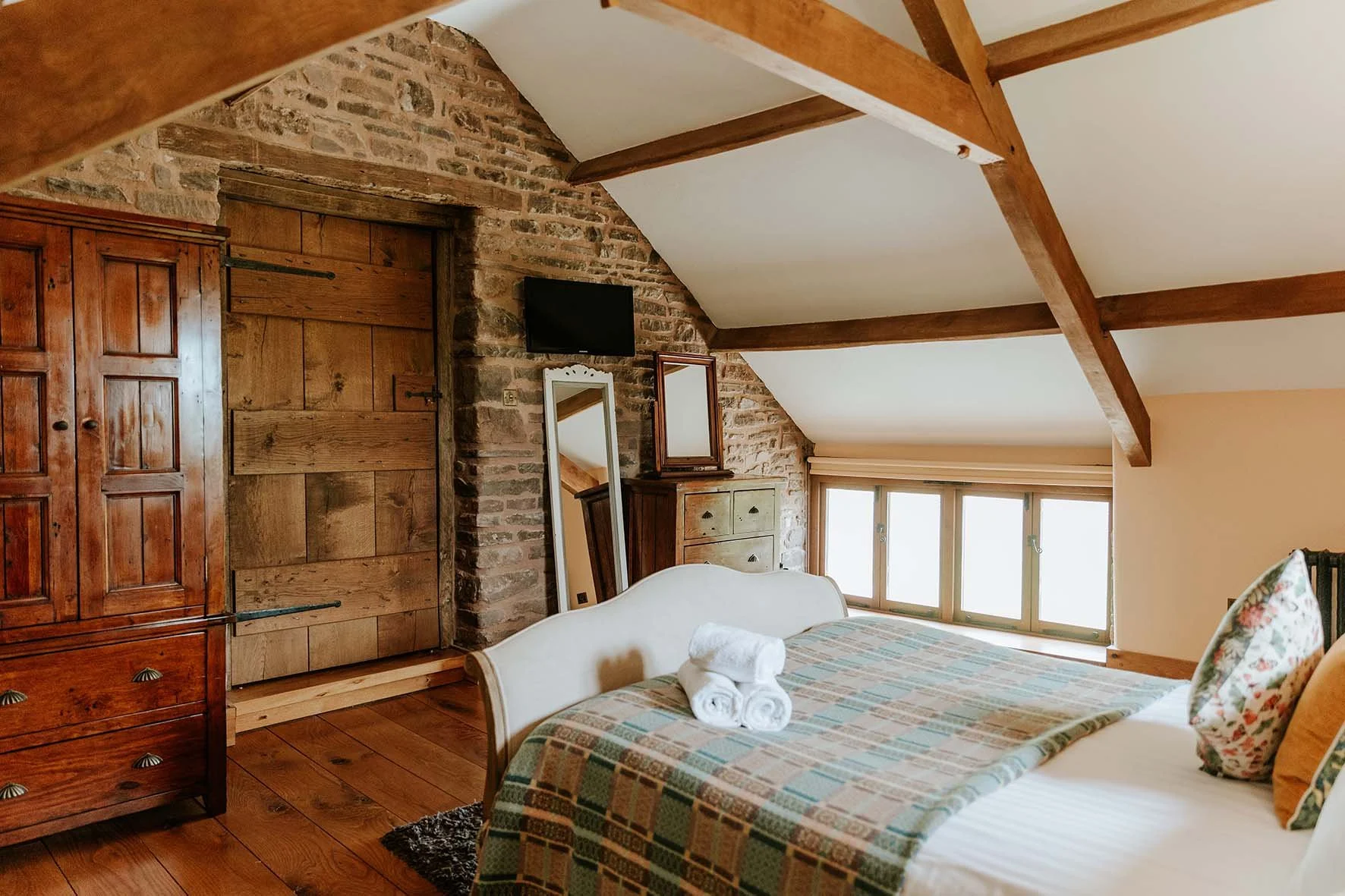 Cozy bedroom with wooden beams, brick wall, and a large window. A bed with patterned blanket and pillows, with towels on top. There is a wooden wardrobe, small dresser, mirrors, and a small mounted TV.