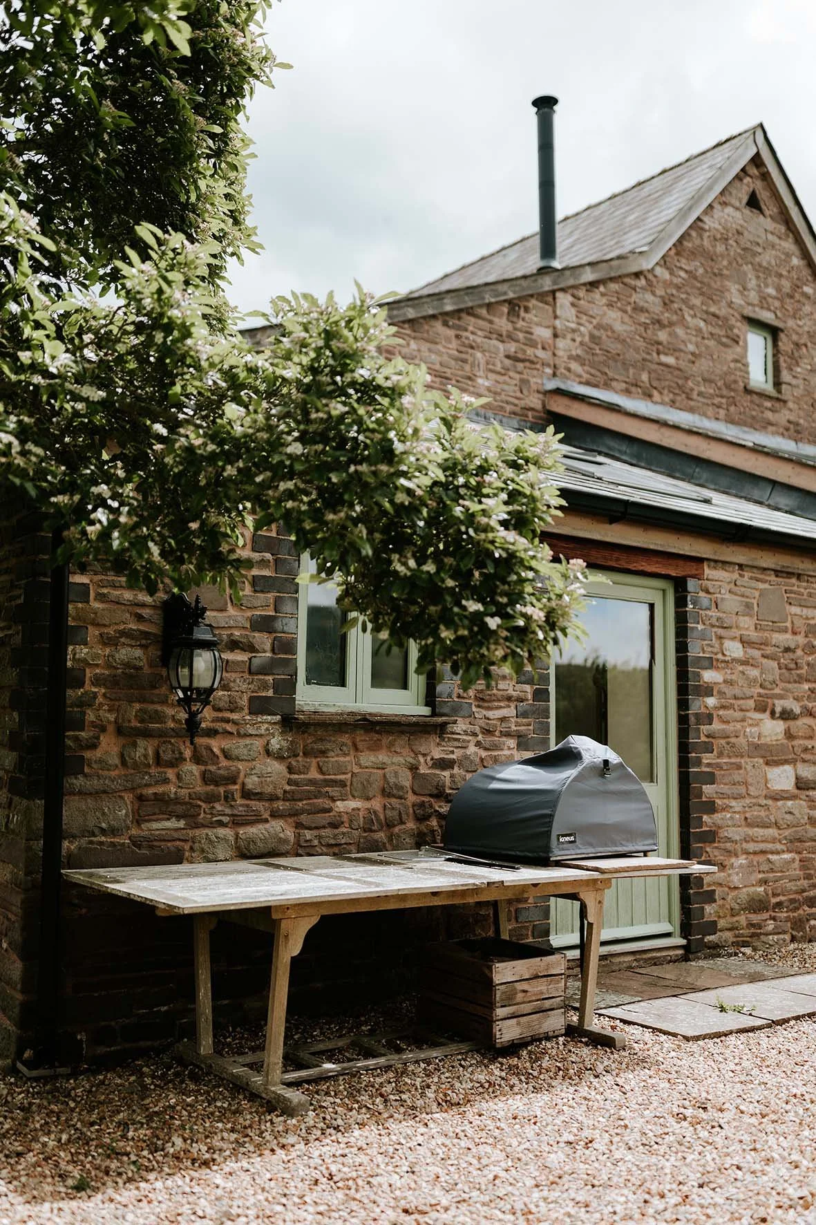 A rustic brick house exterior with a window, a door with a green frame, and a black barbecue grill on a wooden table. There is a flowering tree partially covering the scene and a gravel ground.