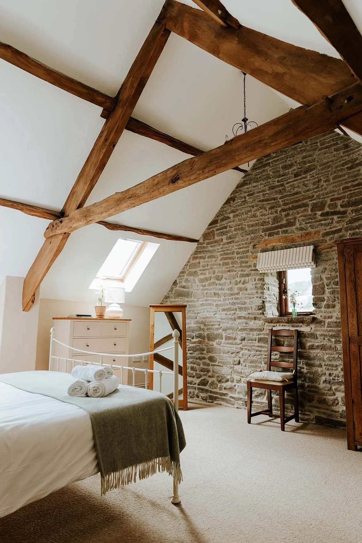 A cozy bedroom with a white metal bed, rolled towels on the bed, a beige carpet, a wooden dresser with a potted plant, a small window with a striped curtain, a wooden chair, a mirror, a skylight, exposed wooden beams, and a stone brick wall.
