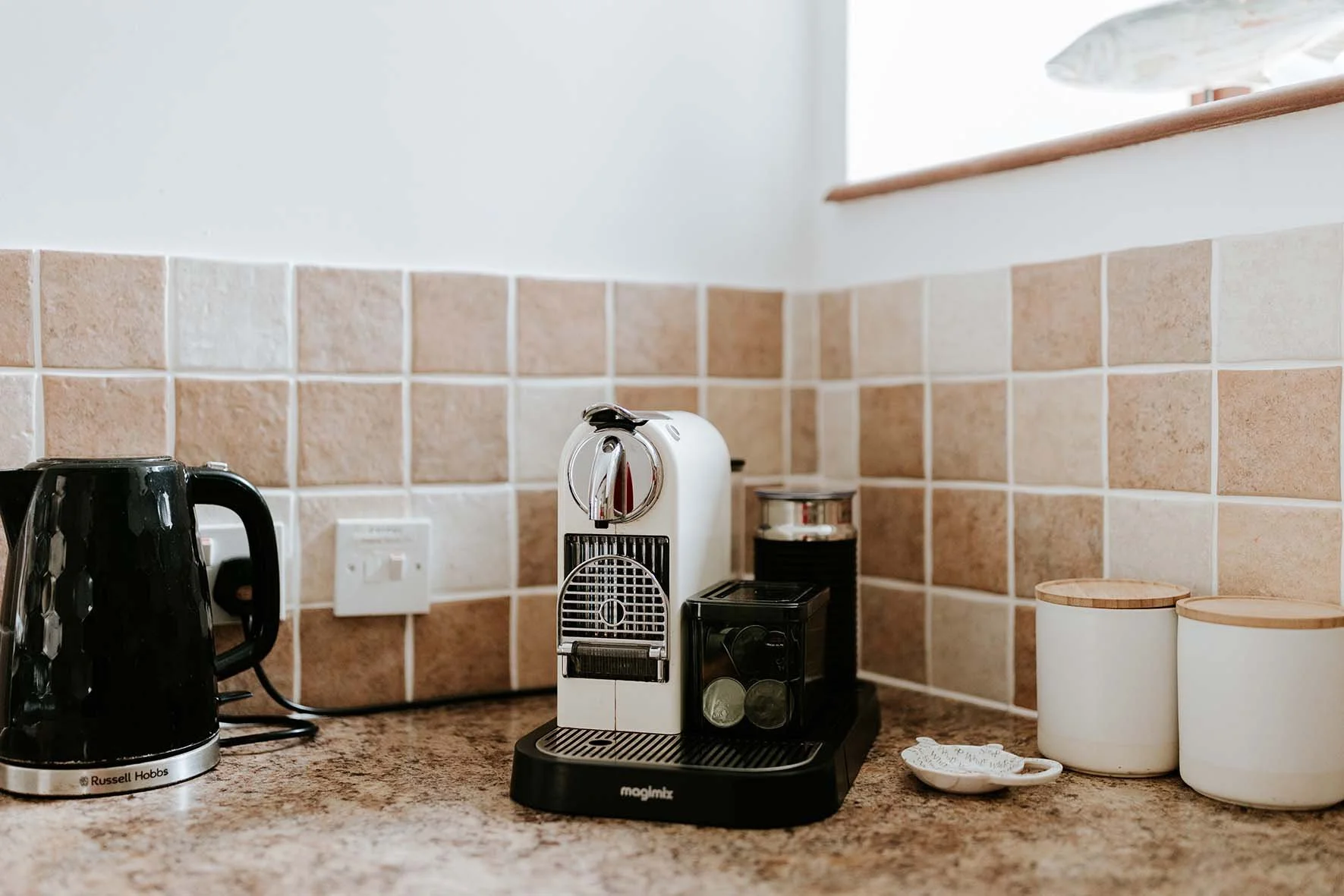 Coffee machine with a black base, a white body, and a silver spout on a kitchen counter with beige tile backsplash, black electric kettle on the left, and two white ceramic containers with wooden lids on the right.