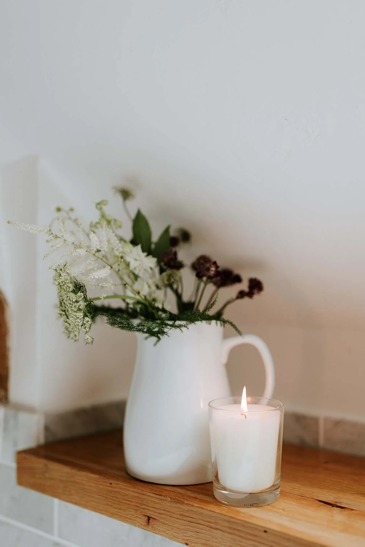 A white ceramic pitcher with flowers next to a lit white candle in a glass holder on a wooden surface, set against a cream-colored wall.