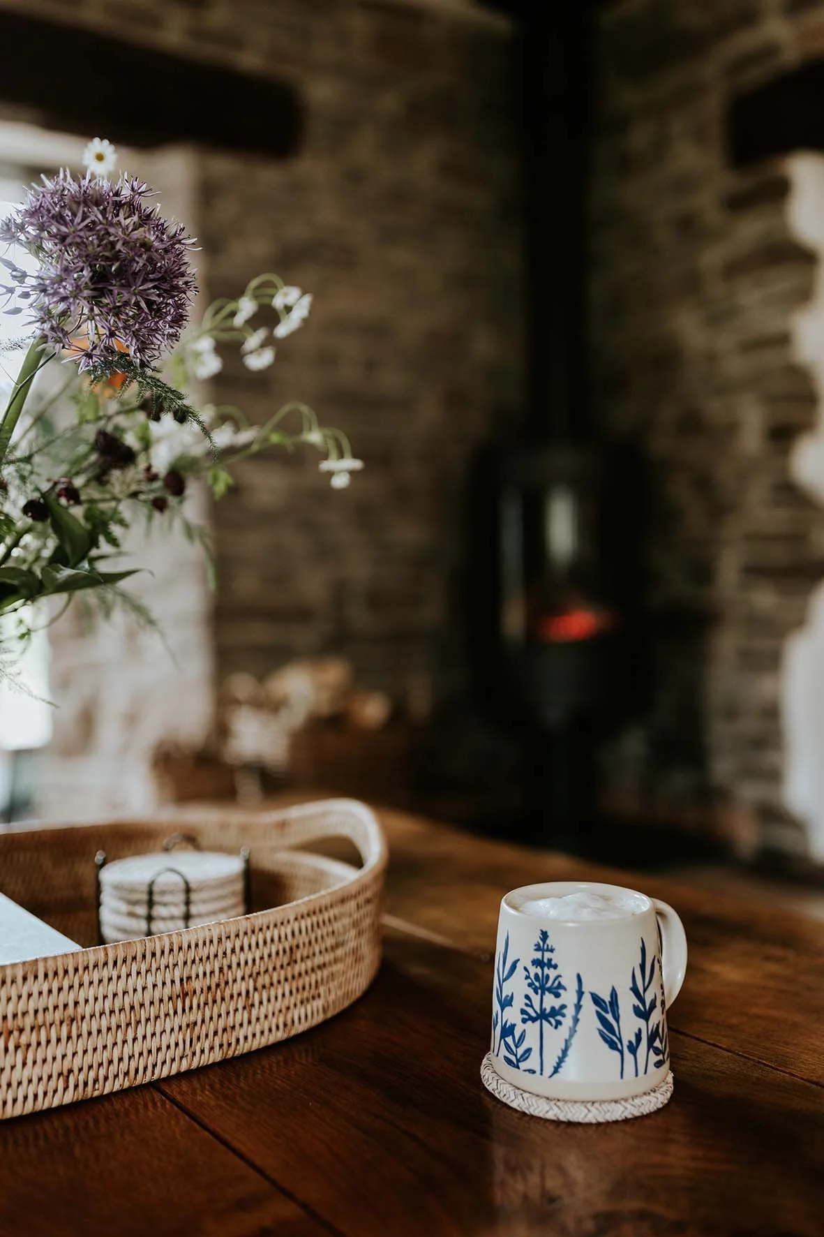 A cozy indoor scene with a wooden table, a mug with blue botanical patterns, a wicker tray, and a bouquet of flowers, against a rustic brick wall with a black stove in the background.