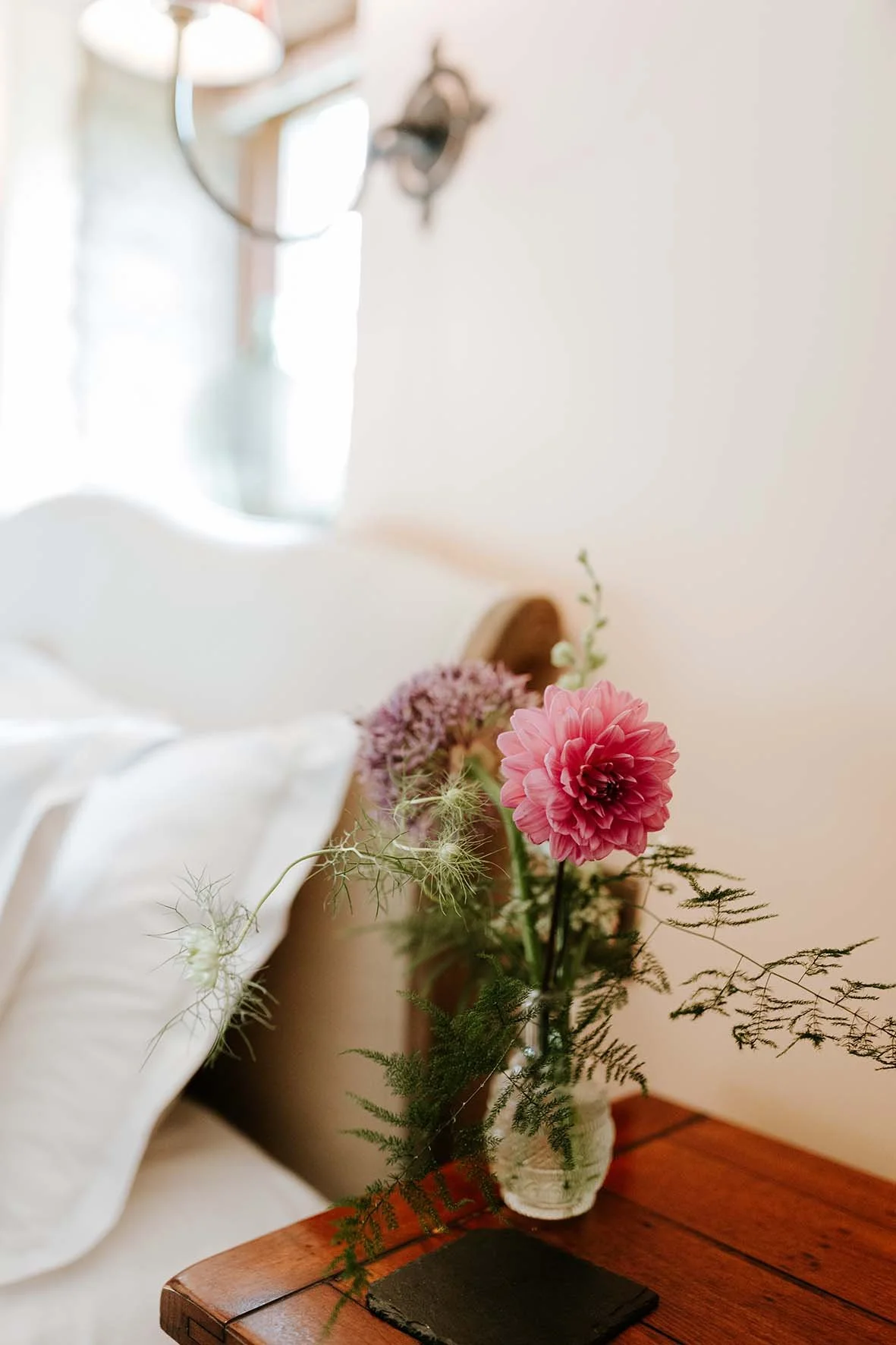 A vase with pink and purple flowers on a wooden table near a bed with white linens in a bedroom with a wall mirror and soft lighting.