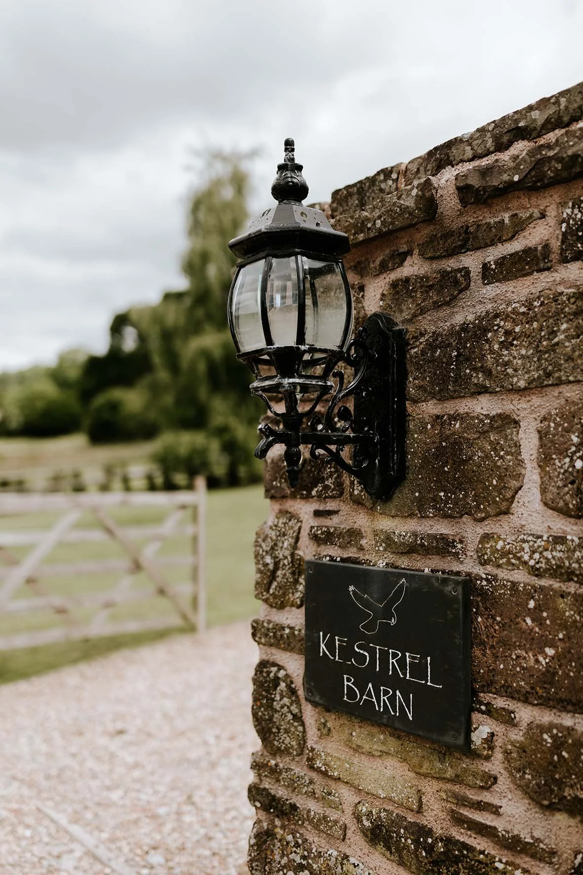 A black outdoor wall-mounted lantern attached to a brick wall, with a black sign below reading "Kestrel Barn" and a minimal outline illustration of a kestrel bird above the text. The background features a fenced outdoor area with green grass and tree