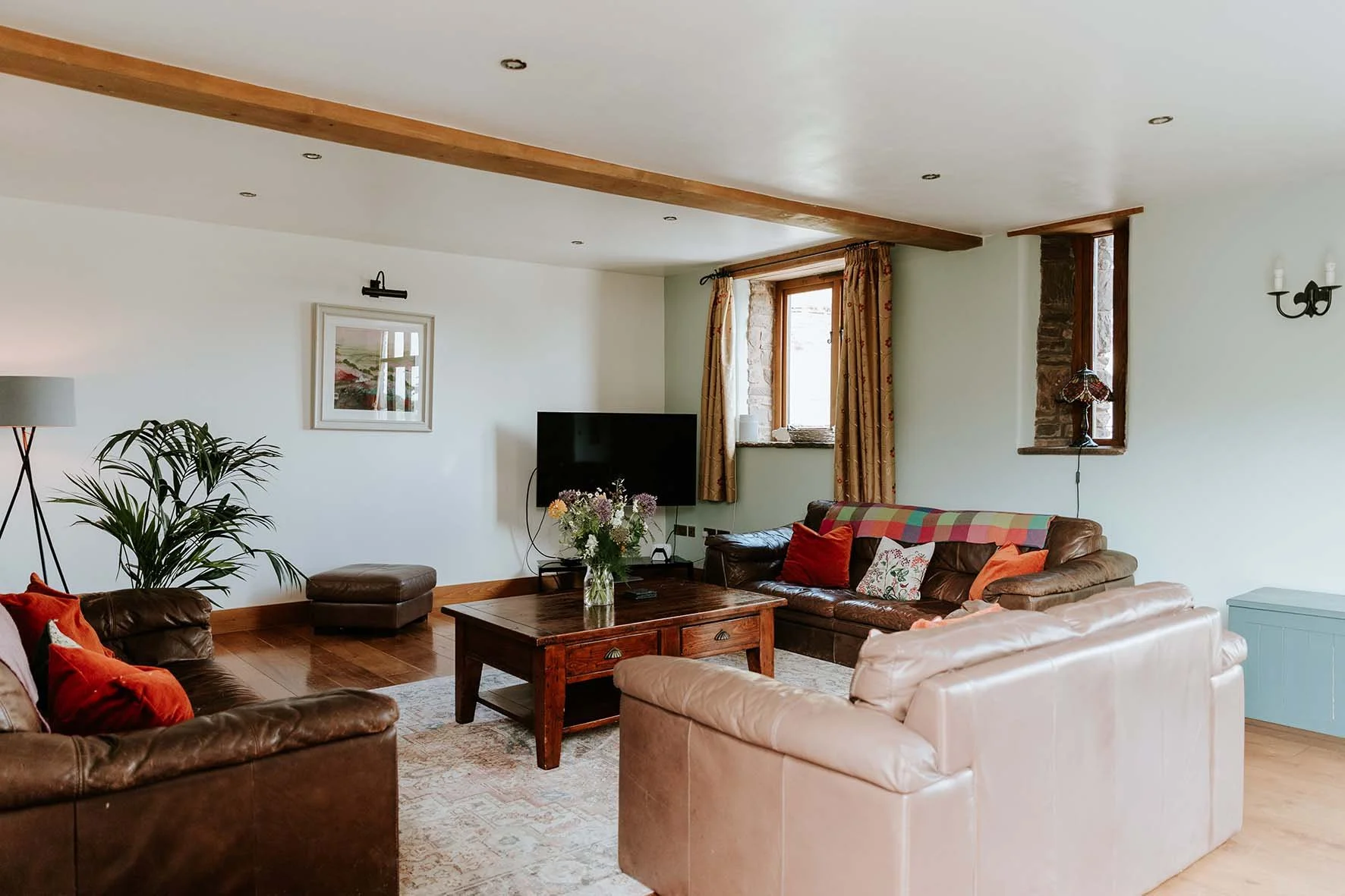 Living room with leather and fabric sofas, a wooden coffee table with a vase of flowers, a flat-screen TV, brick and wooden window frames, curtains, a wall lamp, a tall floor lamp, and framed artwork on the white walls.
