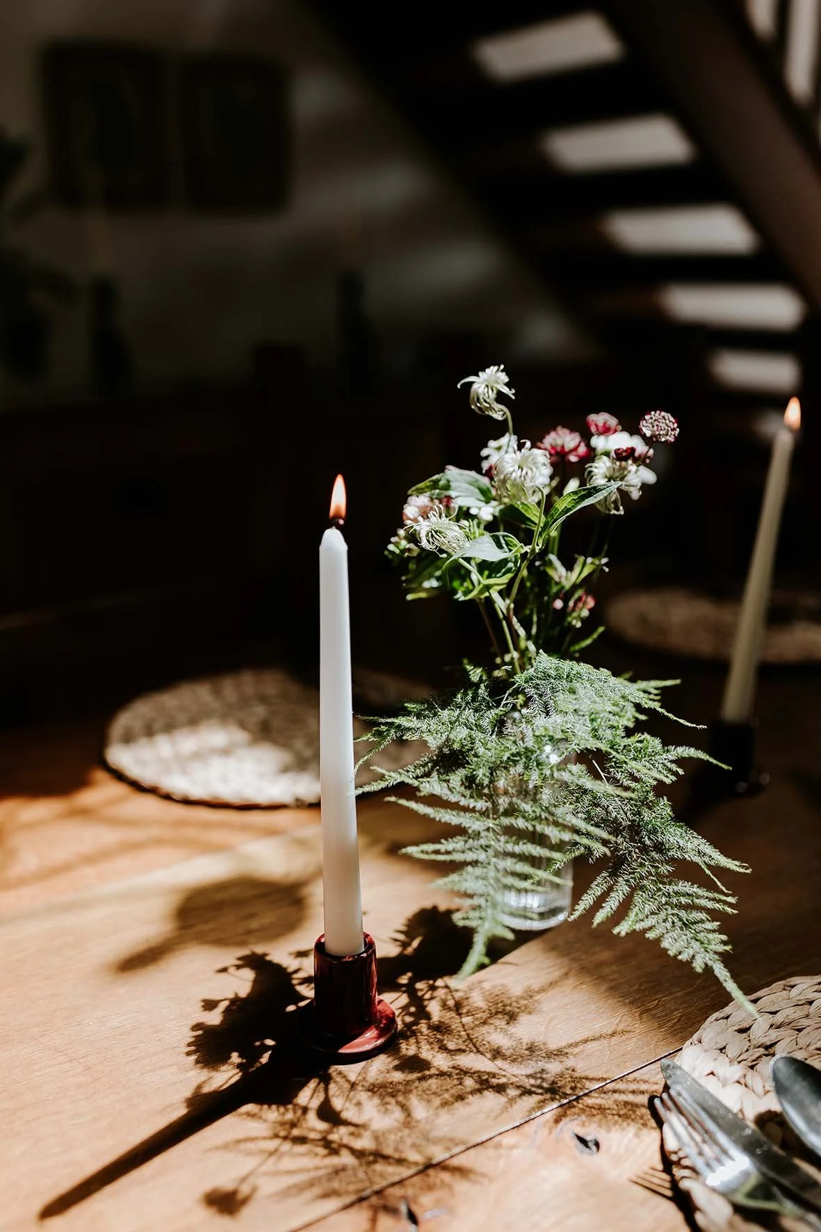 A table with a lit white candle in a red holder and a glass vase containing assorted flowers and greenery. Shadows of the candle and flowers are cast on the wooden table.