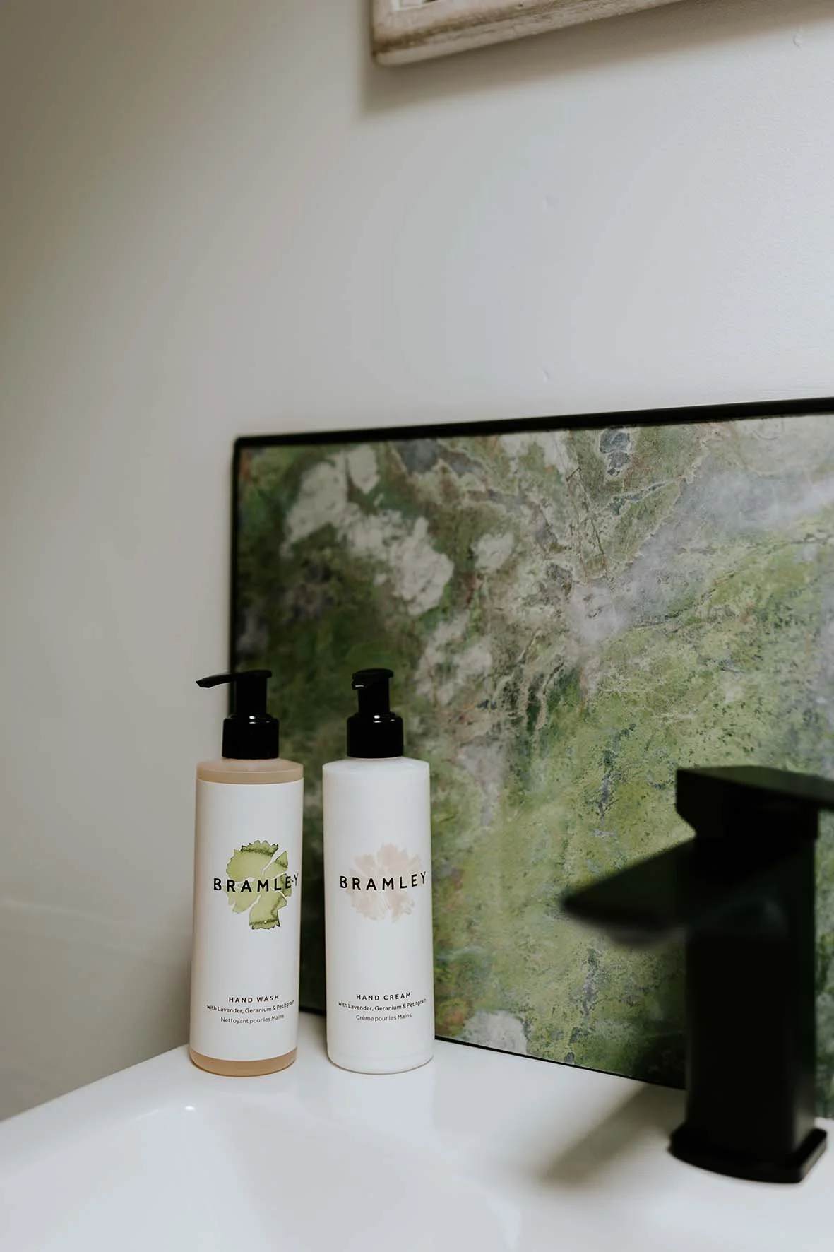 Two Bramley hand soap and hand cream bottles with pump dispensers on a bathroom sink, behind a green and gray marble-patterned backsplash.