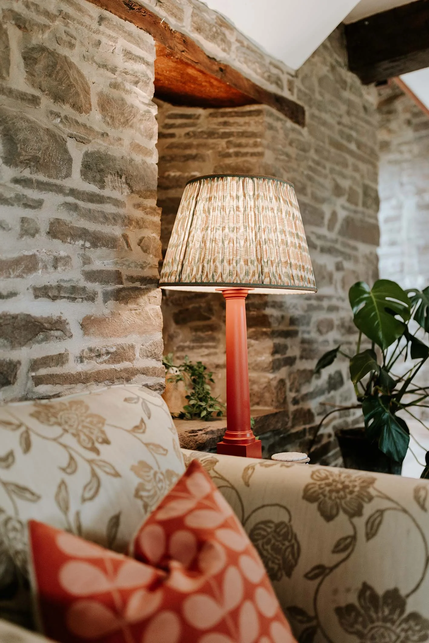 A cozy living room corner featuring a floral-patterned couch, a red table lamp with a patterned shade, a potted plant, and an exposed brick wall with wooden beams.