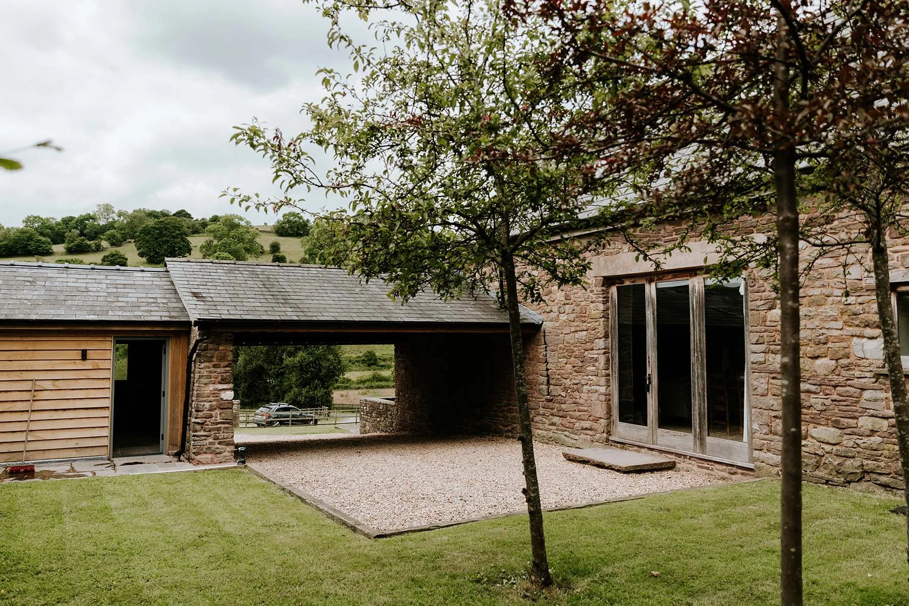 View of a rustic stone house with a gravel patio, surrounded by a grassy lawn and trees, with hills in the background under cloudy skies.