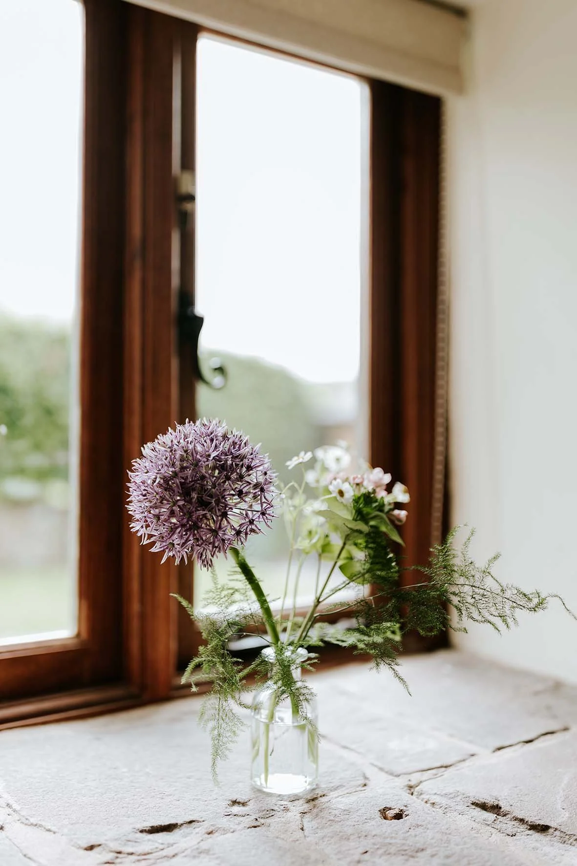 A vase with a bouquet of flowers on a windowsill, with a wooden window frame and a blurry outdoor scene visible through the window.