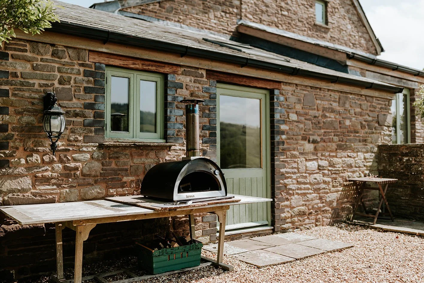 A rustic brick house with light green window frames and a door. Outside, there is a wooden table with a portable pizza oven on top, a lantern attached to the wall, and a small foldable table nearby. The ground is covered with gravel and stone slabs.