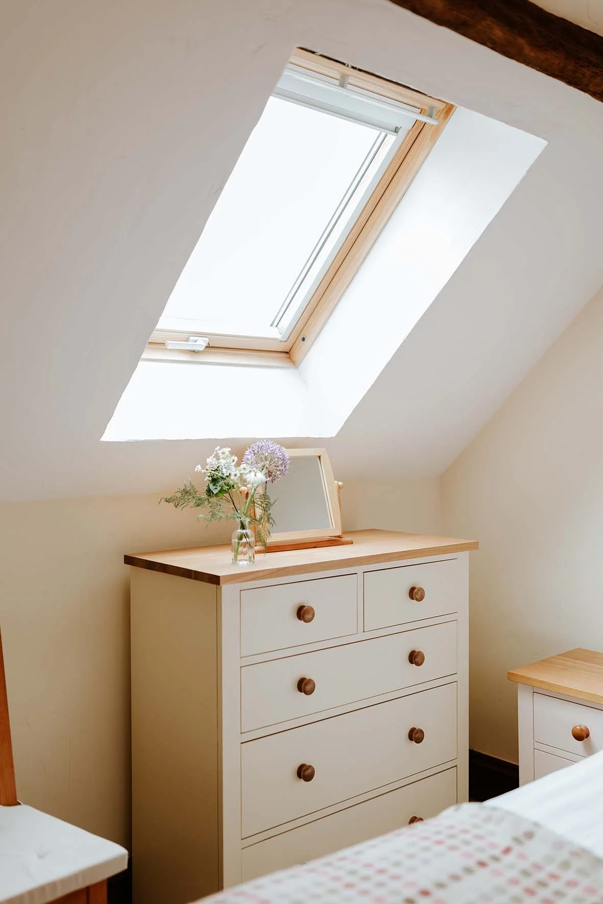 A white dresser with wooden knobs, topped with a glass jar of flowers and a small mirror, beneath a skylight window in a bedroom with beige walls.