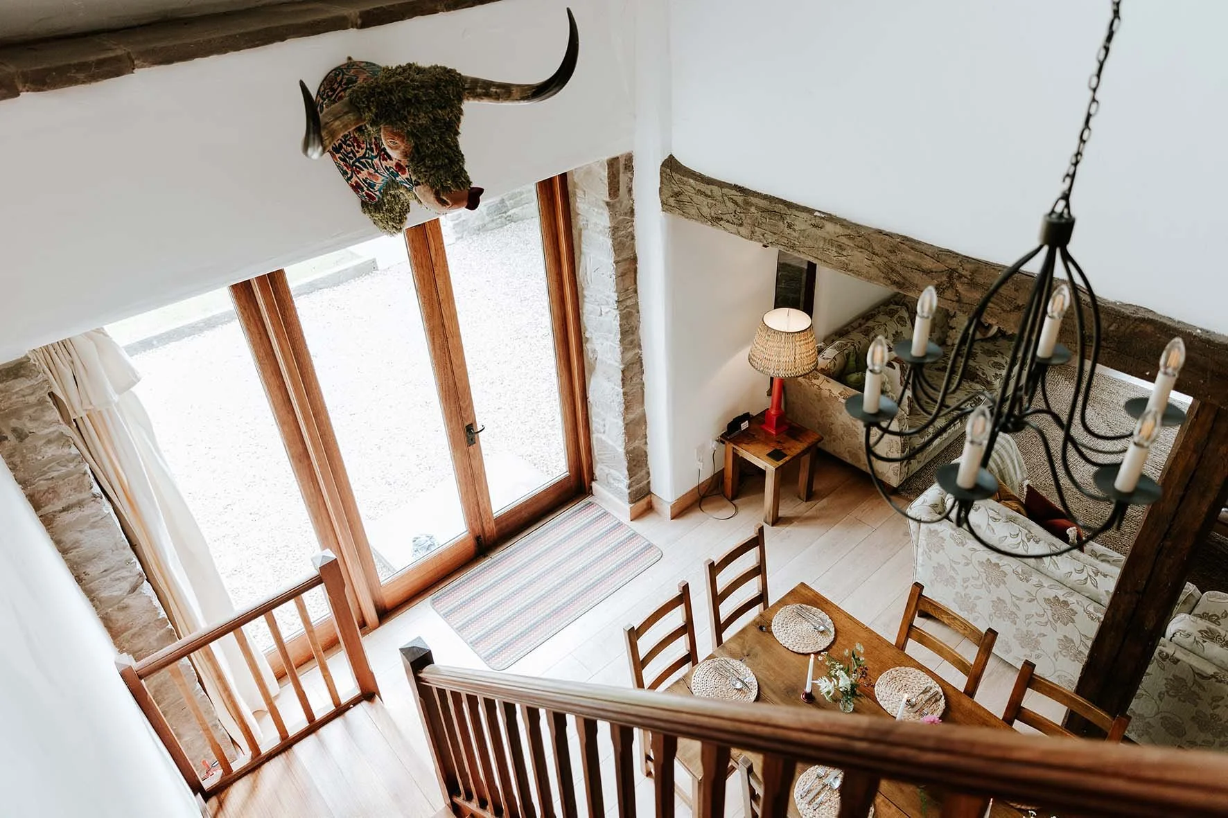 An aerial view of a living room and dining area with wooden furniture, a chandelier, a large glass sliding door, a staircase, a sofa, a lamp, and a decorative buffalo head with horns on the wall.