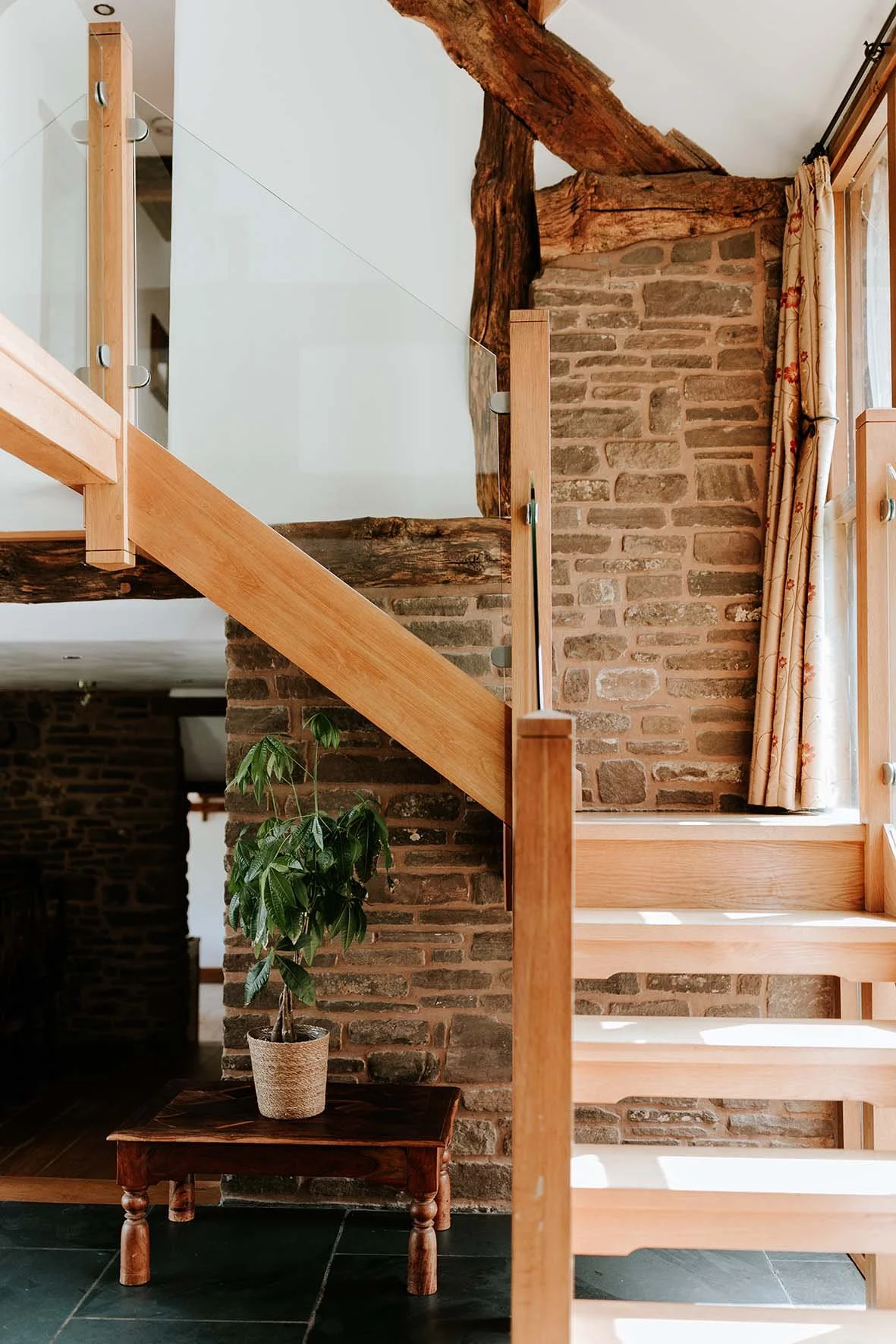 Interior view of a staircase with wooden steps, brick wall, exposed wooden beams, a potted plant on a small wooden table, and a window with floral curtains.