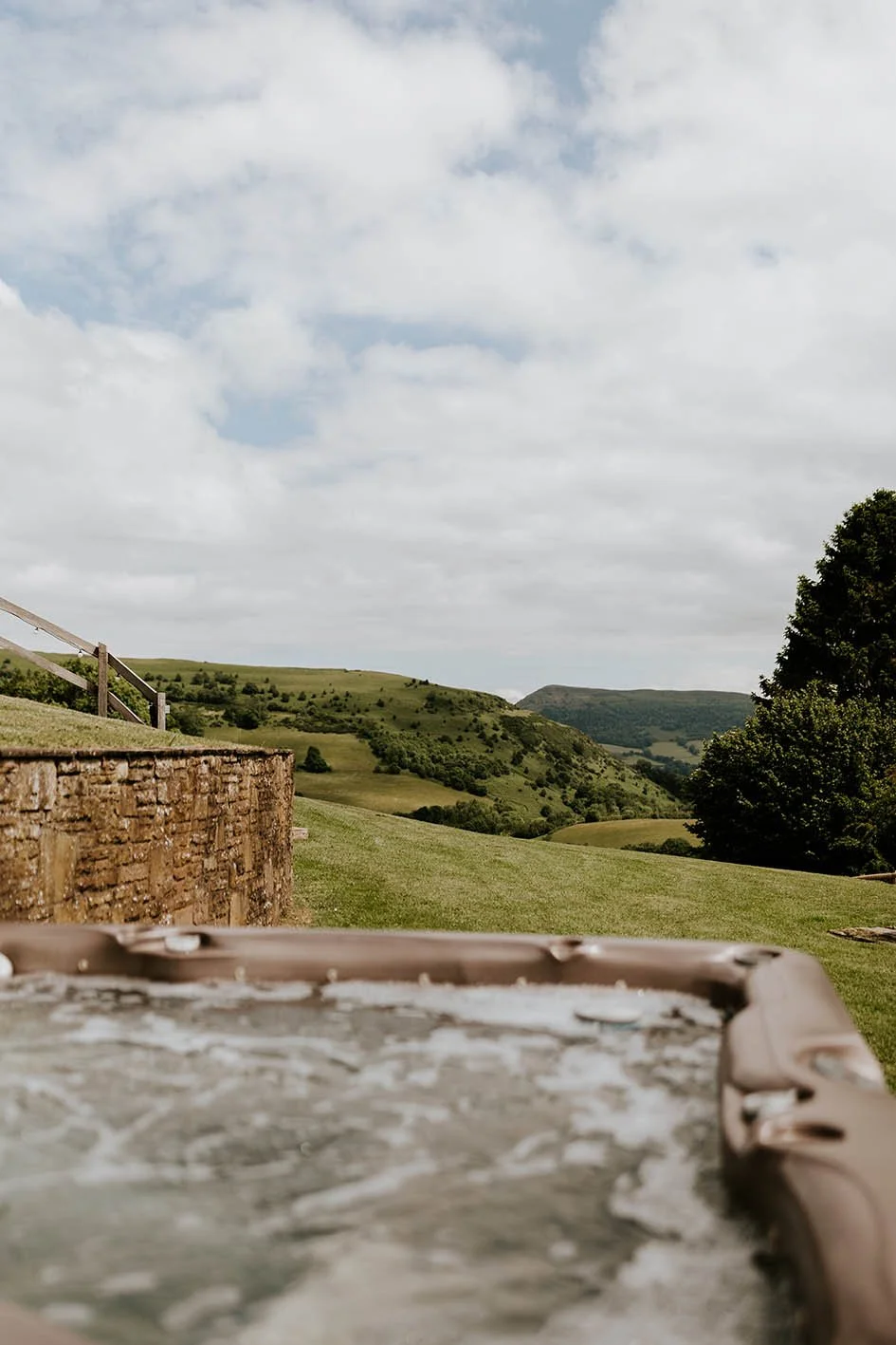 A scenic view of rolling green hills with a cloudy sky, a stone wall, and part of a hot tub in the foreground.
