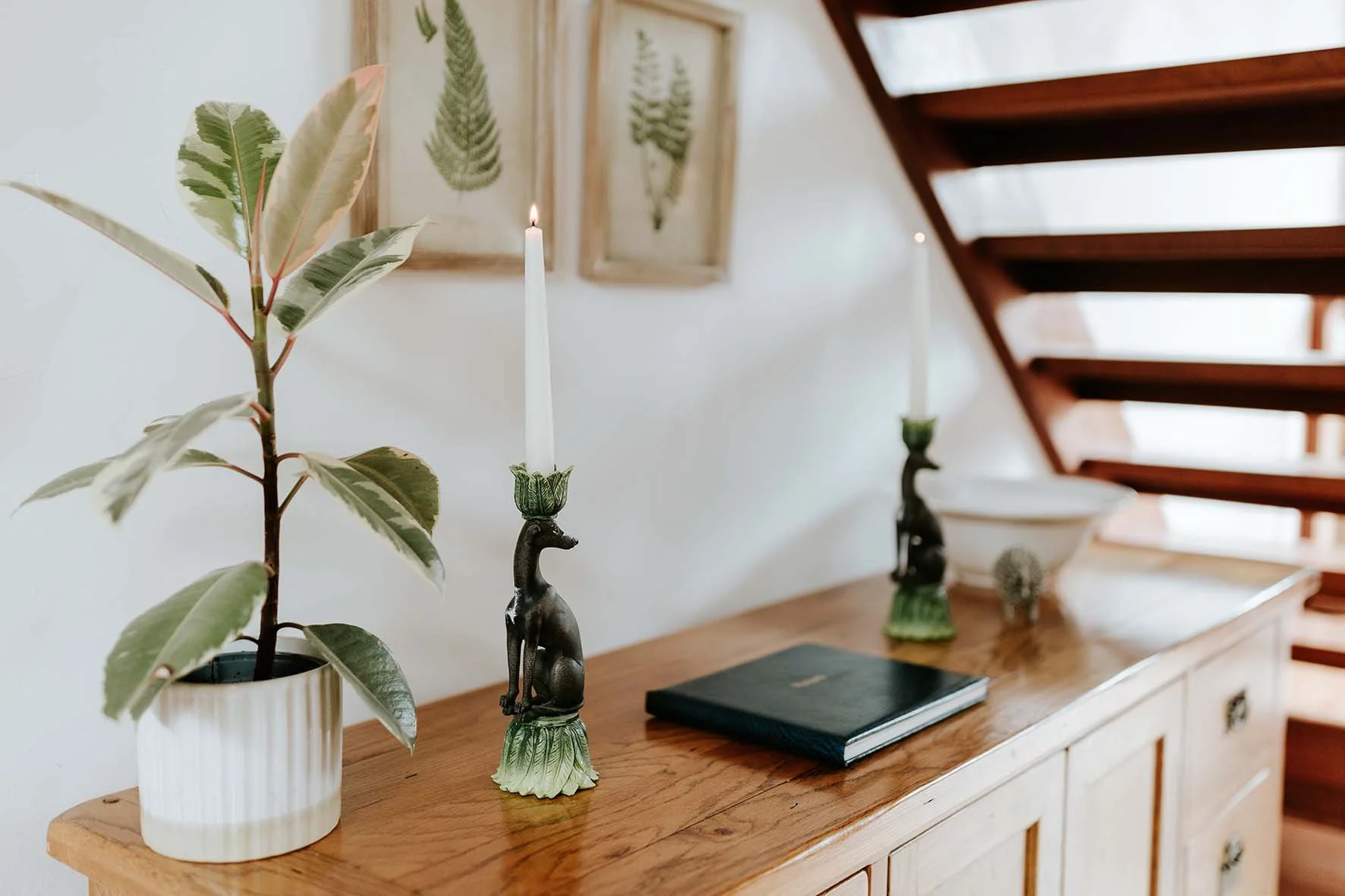 A wooden sideboard with decorative items including a potted plant, two ornate candle holders with white candles, a closed black book, a small decorative globe, and framed botanical art on the wall, next to a staircase with wooden steps.