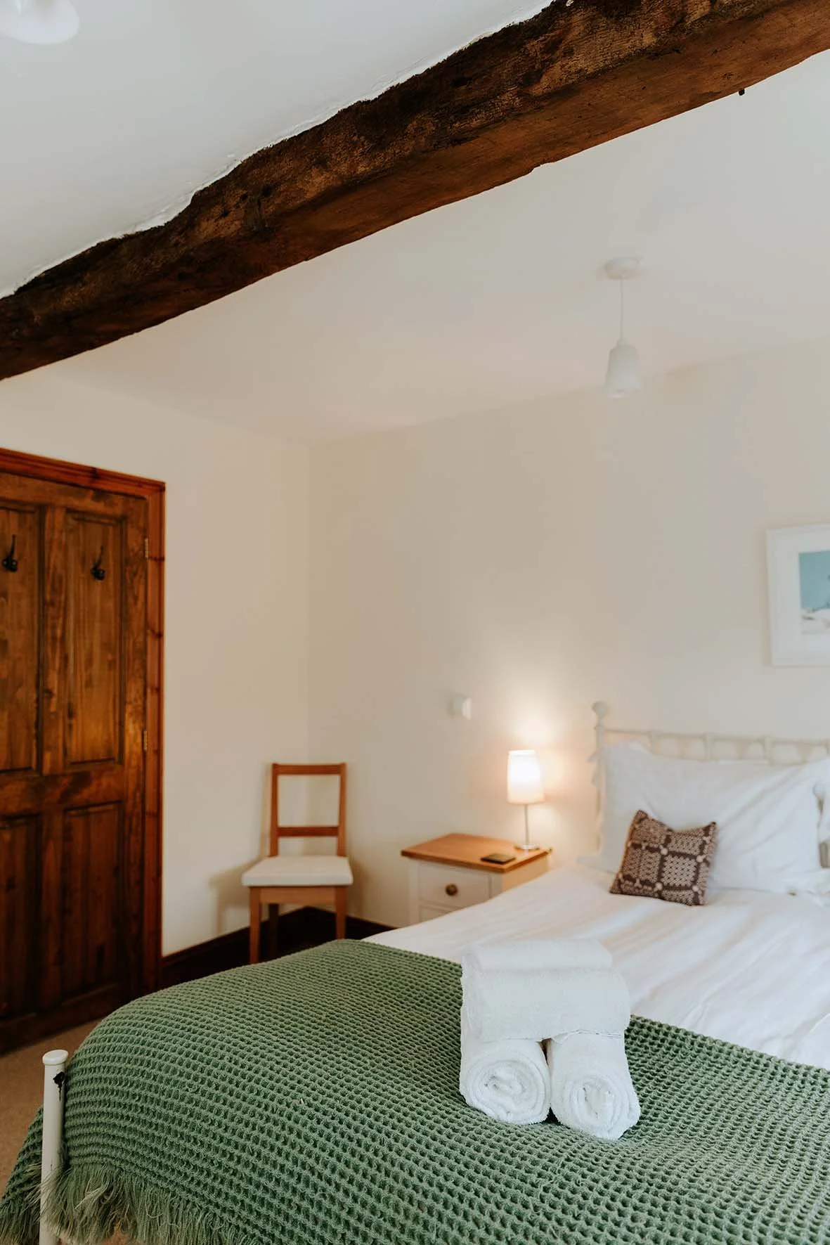 A cozy bedroom with a wooden beam on the ceiling, white walls, a bed with white bedding, a green textured blanket at the foot of the bed, and two rolled white towels. A small wooden nightstand with a lamp, a chair with a white cushion, a wooden close