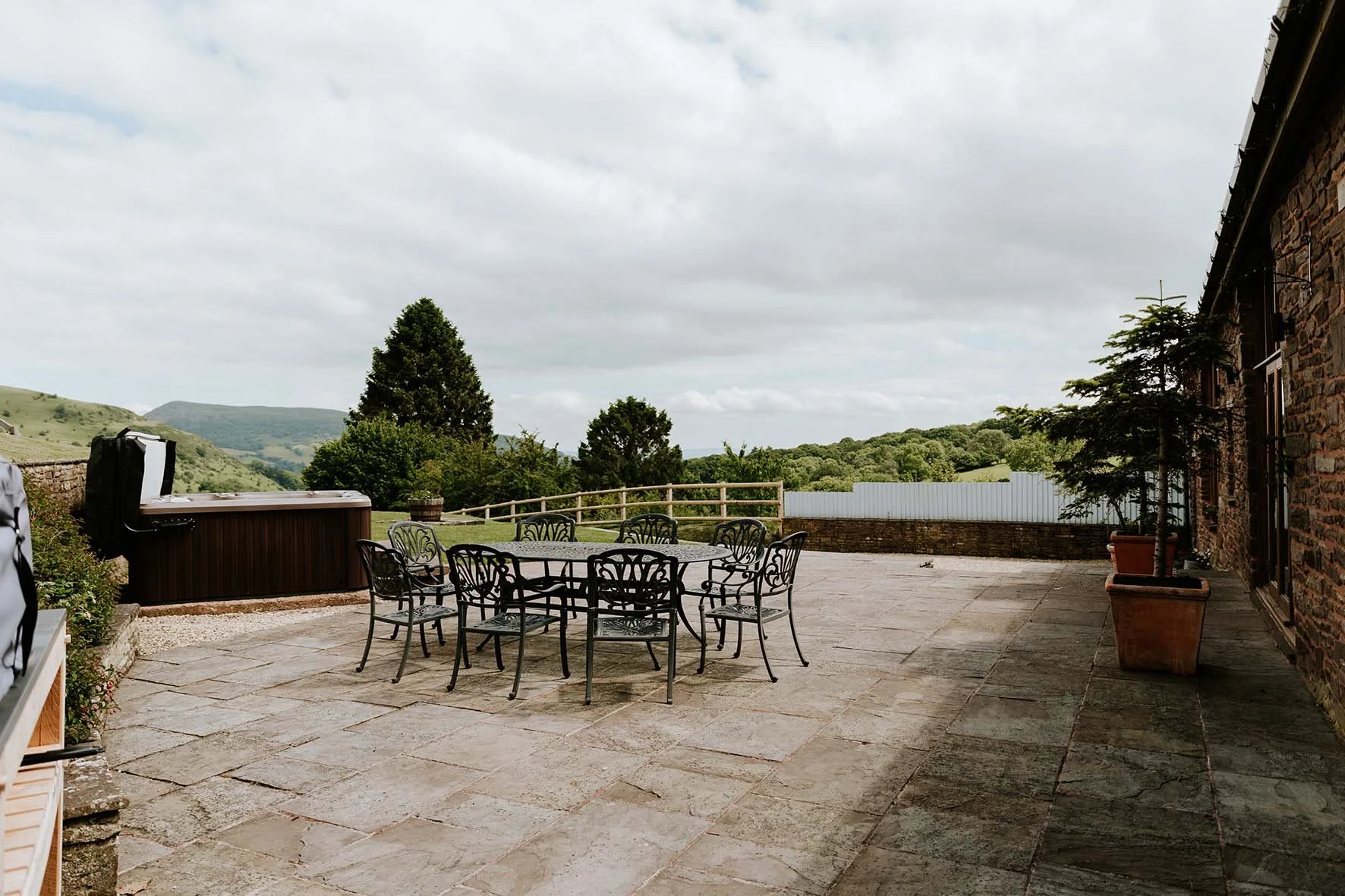 Outdoor patio with a round dining table and chairs, potted plants, a hot tub, and scenic hills in the background.