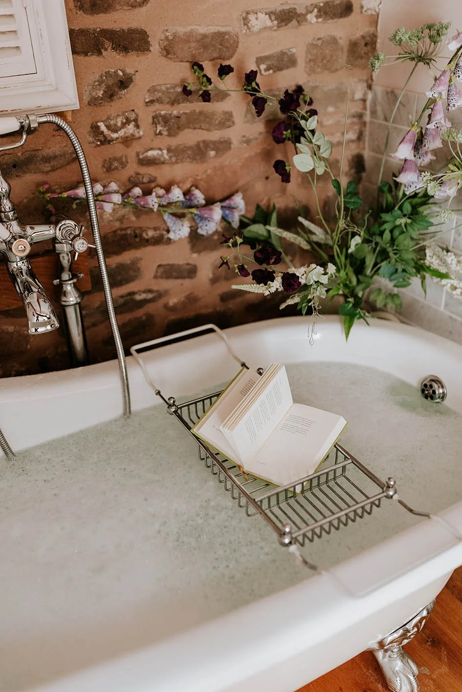 A vintage white clawfoot bathtub filled with water in a bathroom with an exposed brick wall. There is a wire basket with an open book resting across the edge of the tub, and a large arrangement of flowers with purple and green foliage placed beside it.