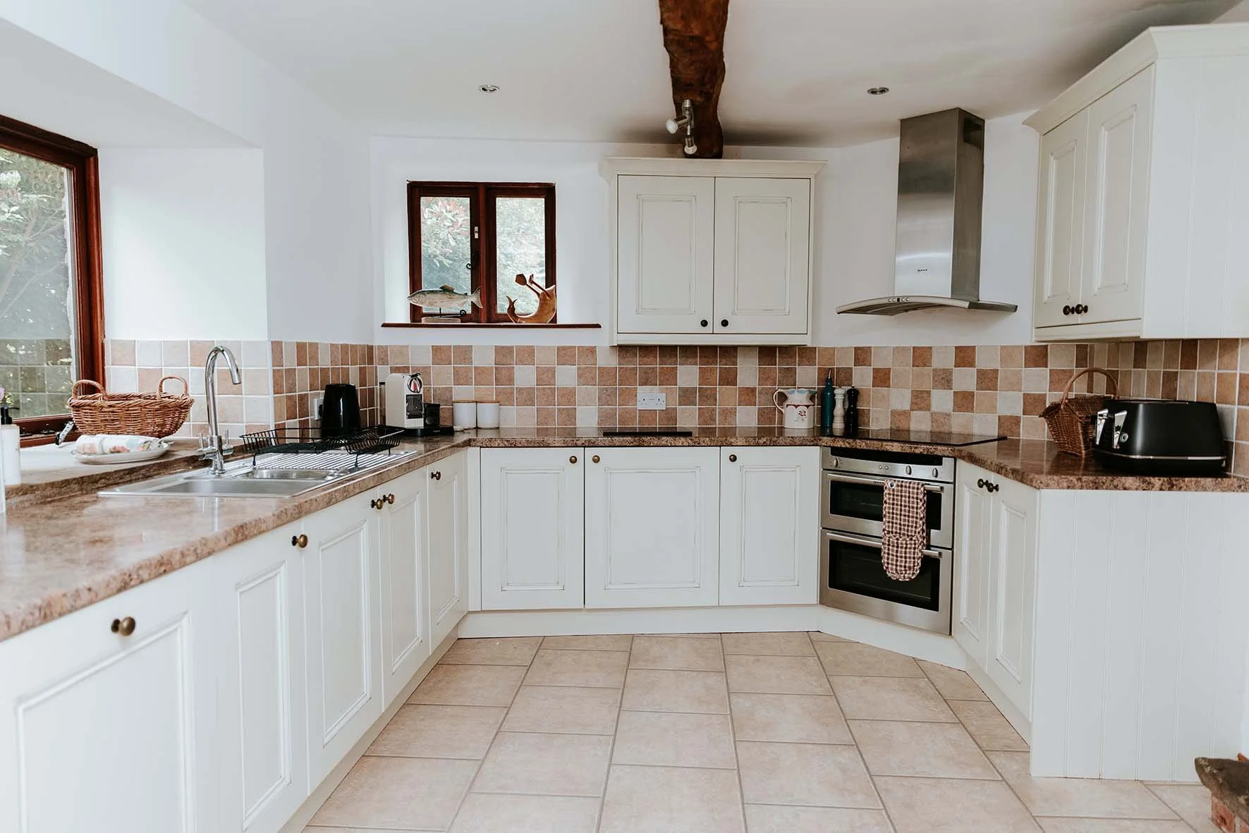 A bright, clean kitchen with white cabinets, beige tiled backsplash, and brown granite countertops. Features include a double sink, small window, and various appliances such as a black kettle, coffee machine, toaster, and oven. There is a decorative 