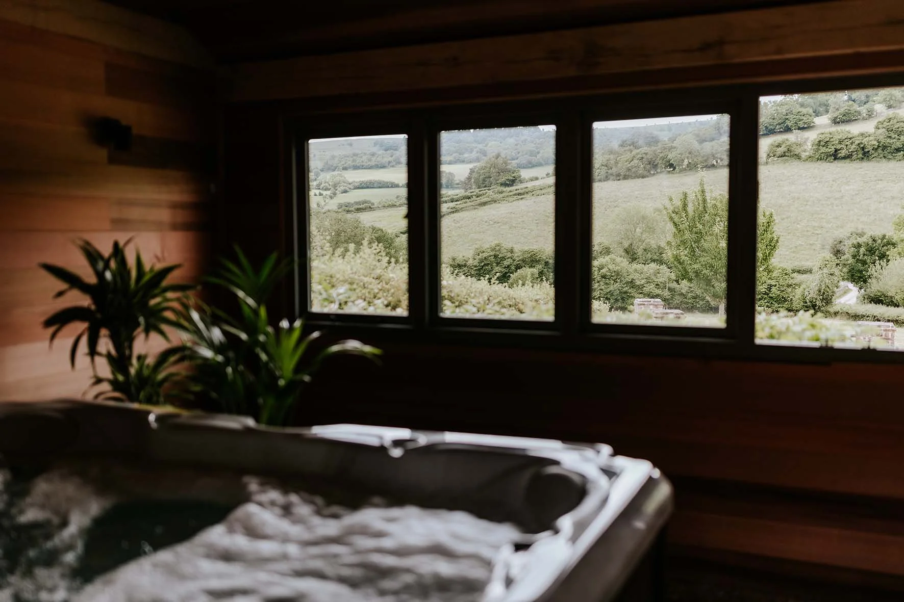 Interior of a wooden room with a hot tub near a window showing a scenic view of rolling green hills and trees.