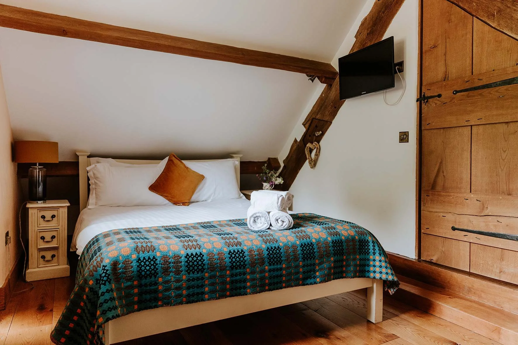 A cozy bedroom with a white bed, orange and white pillows, a small nightstand with a lamp, rolled white towels on the bed, a flat-screen TV on the wall, and wooden beams and panels.